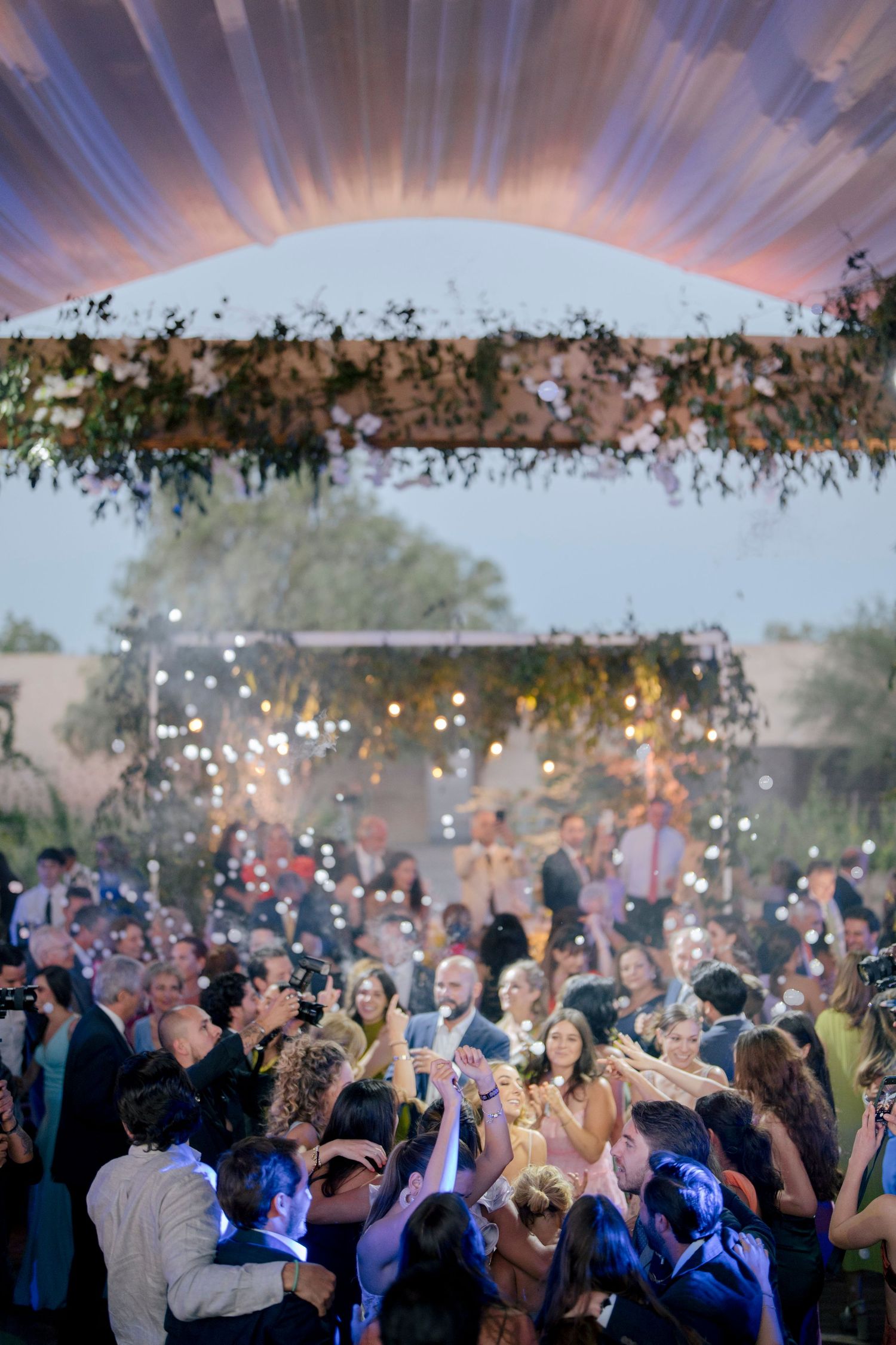 A crowd celebrates at an outdoor event under twinkling lights and a dramatic sunset sky with artistic effects.