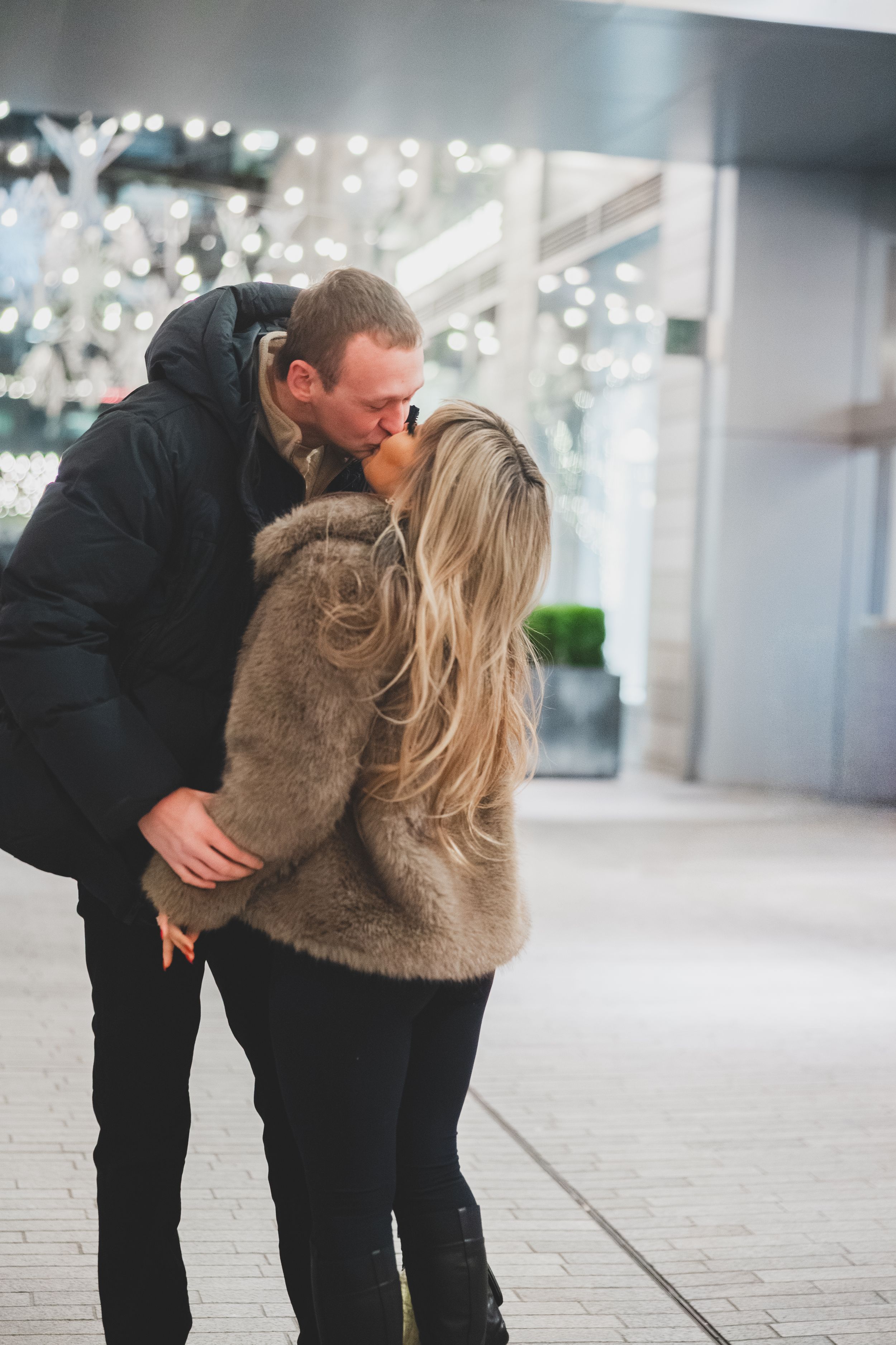 A couple in winter clothing shares a romantic moment outdoors with twinkling lights in the background.