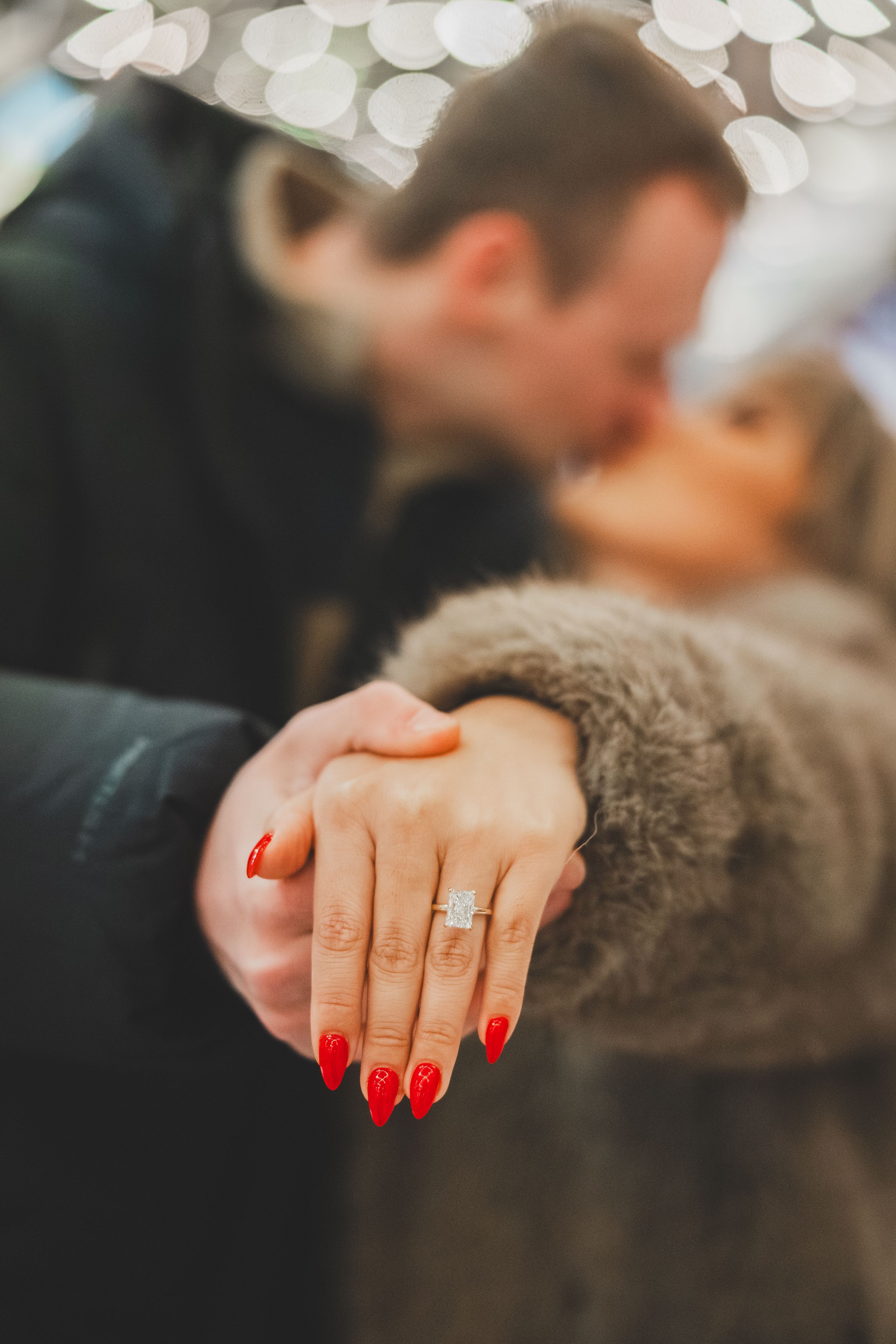 Close-up shot of a hand with bright red manicured nails and a new engagement ring against a blurred background.