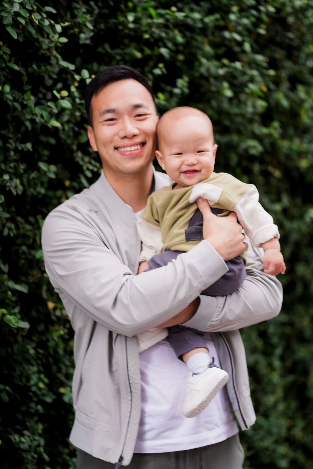 A parent in a gray cardigan holds a baby in beige clothing against a green leafy background.