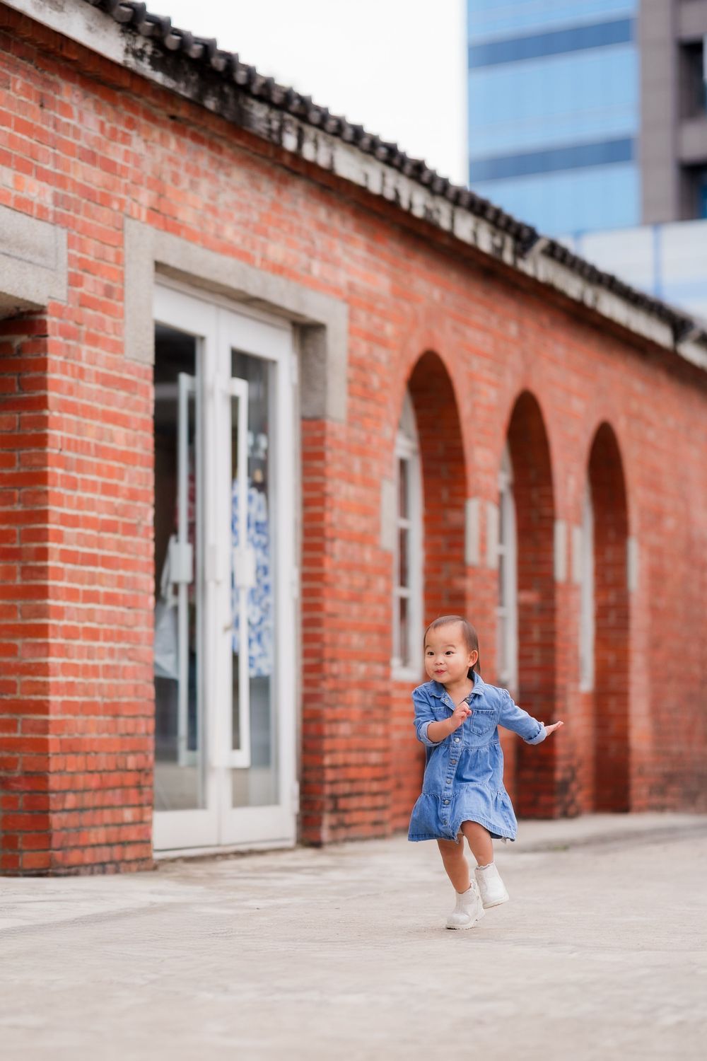 A person in denim sits and poses in front of a red brick building with arched windows and white trim.