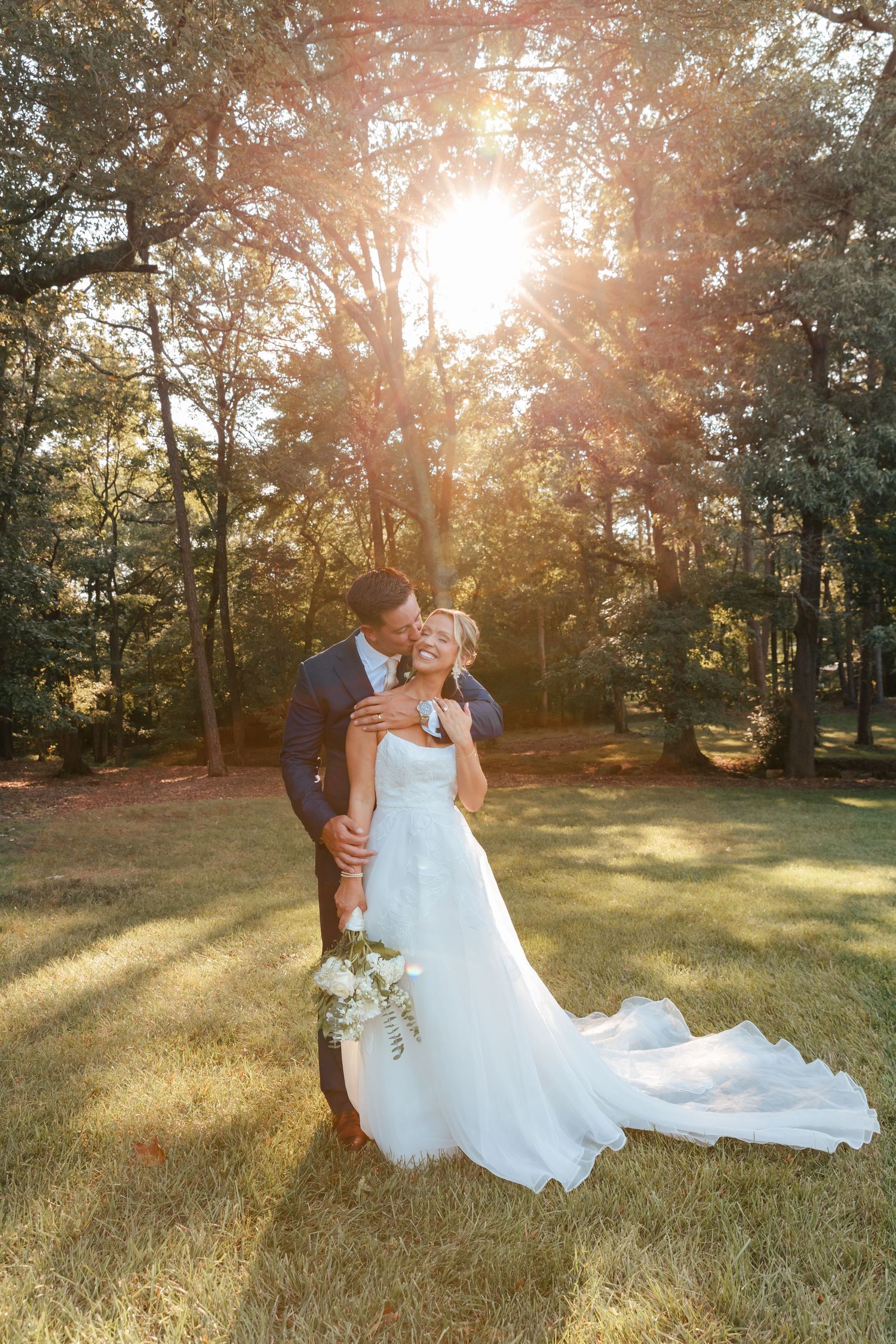 A bride and groom embrace in a sunlit forest clearing during their outdoor wedding portrait.