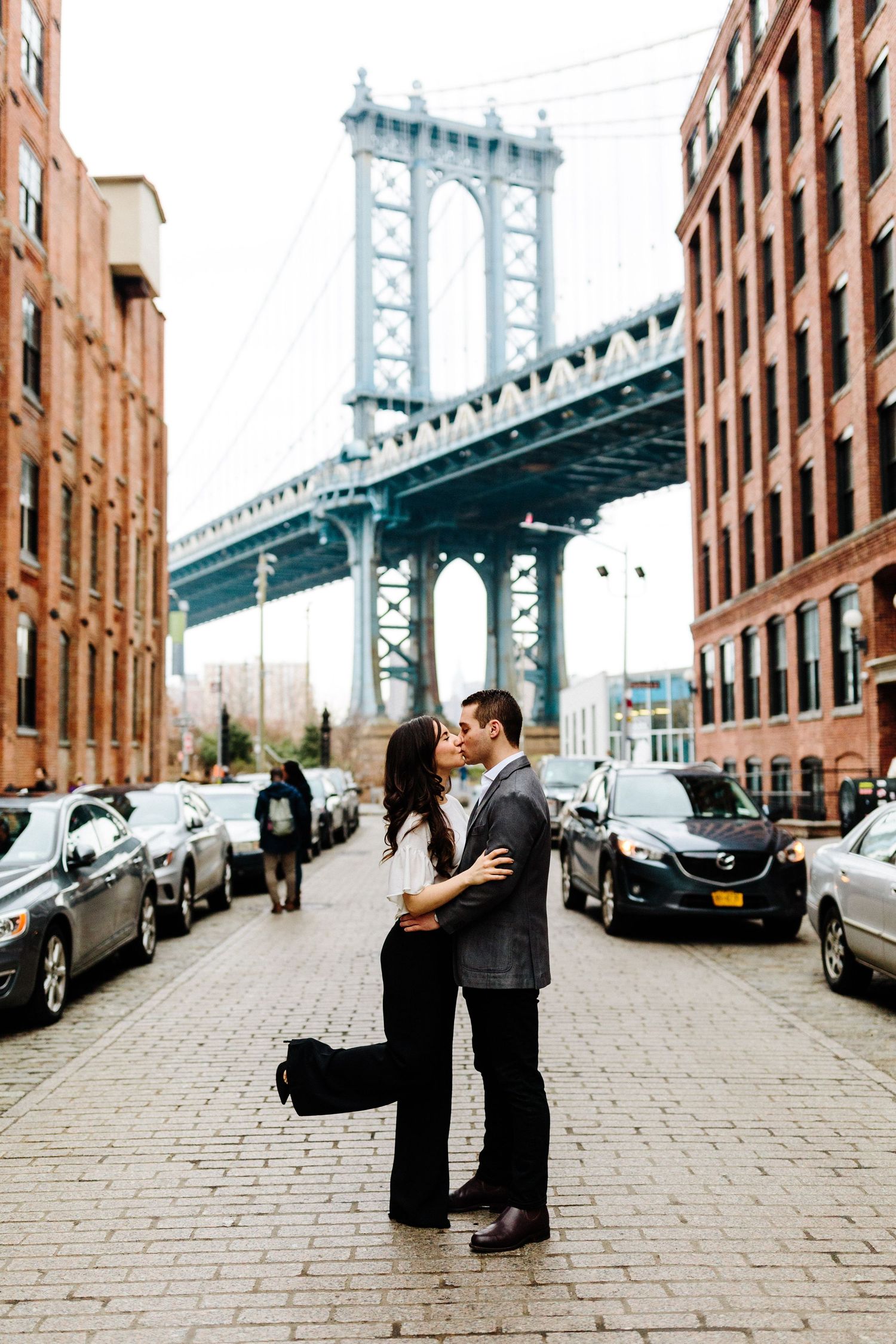 Couple poses on cobblestone street with Manhattan Bridge and brick buildings in background.