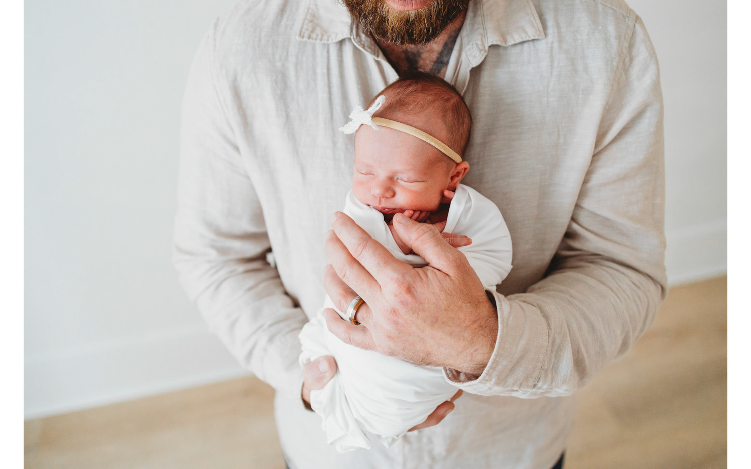 A person in a light colored shirt holds a sleeping newborn baby swaddled in white against a neutral background.