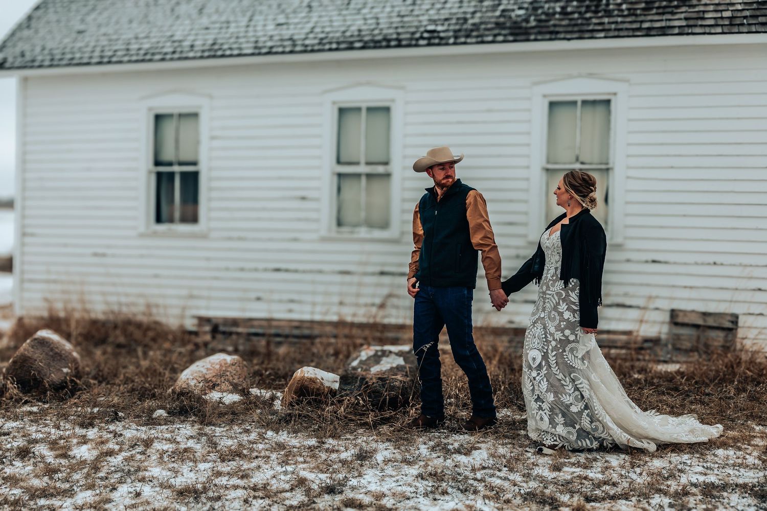 A couple walks hand in hand along a rustic farmhouse exterior in a romantic countryside setting.