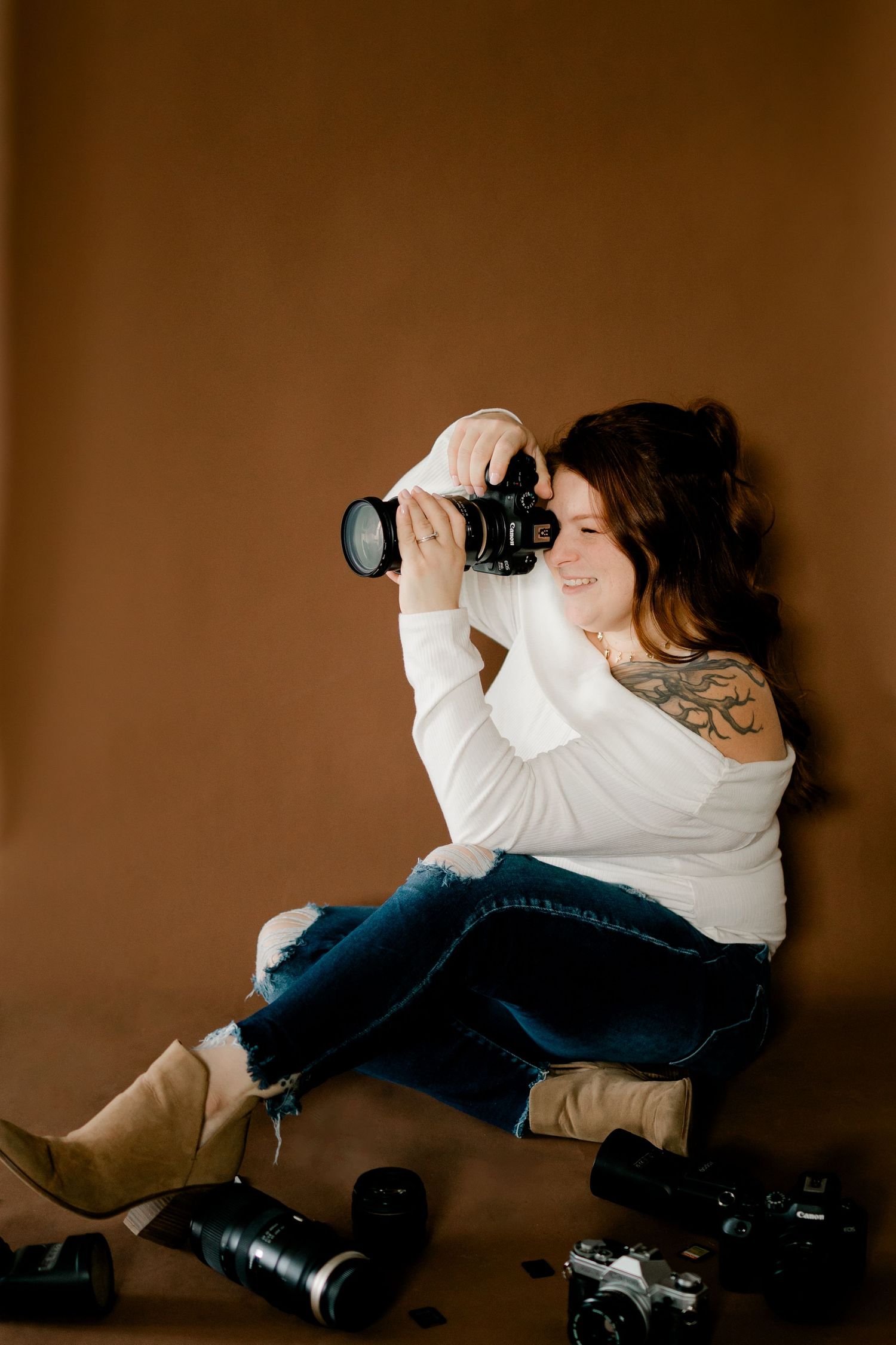 Female photographer sitting on floor holding camera with lenses, wearing white top and jeans.