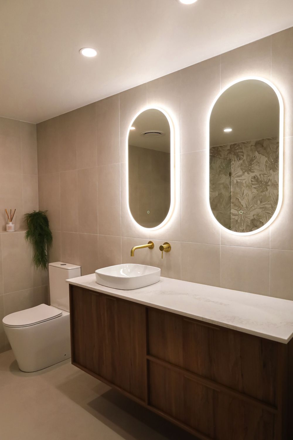Modern bathroom interior with backlit oval mirrors, floating wooden vanity, and vessel sink with gold fixtures.