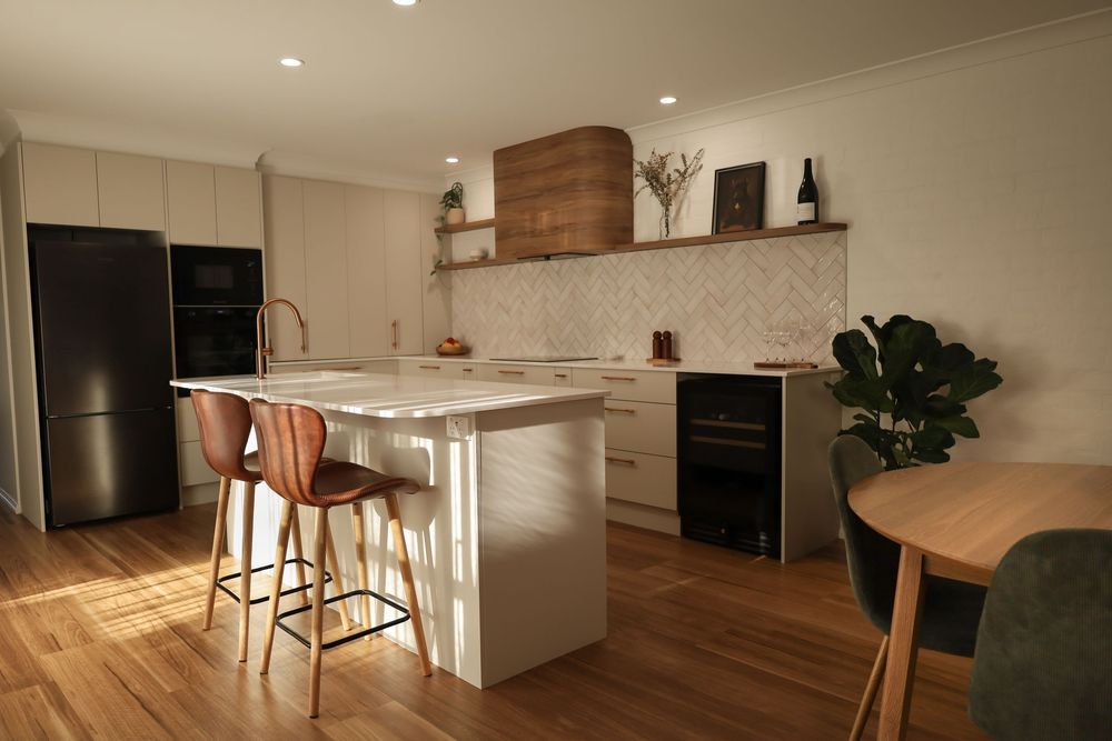 Modern white kitchen with wooden accents features a waterfall island, black appliances, and hardwood flooring.