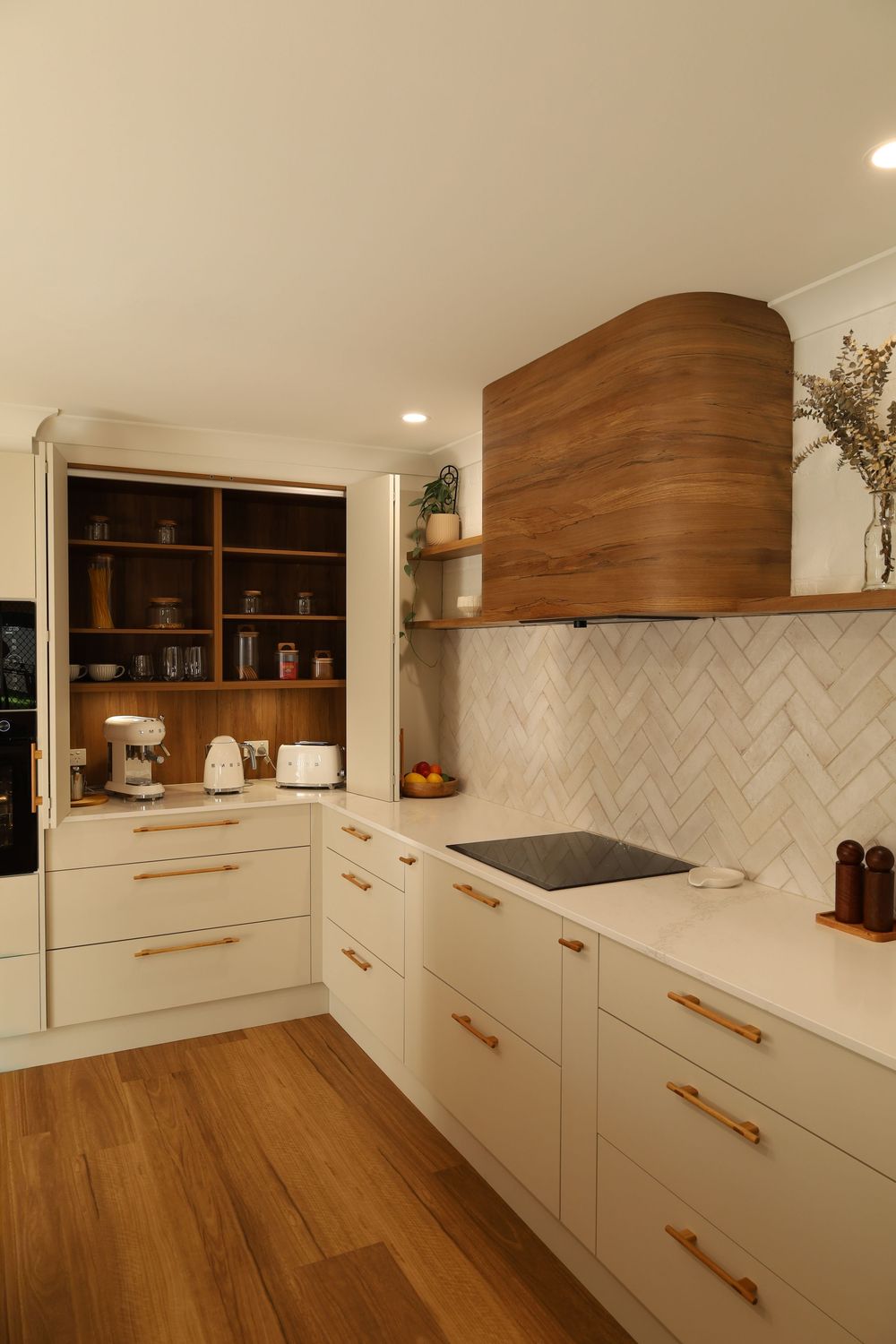 Modern white kitchen with wooden open shelving, copper handles, and herringbone tile backsplash.