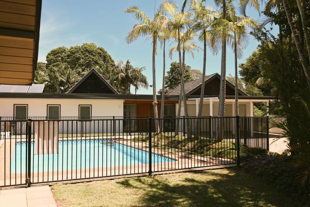 A panoramic view of a backyard swimming pool surrounded by palm trees and black metal fencing.