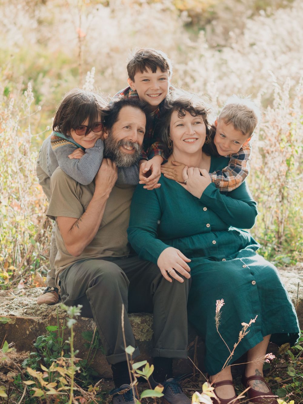 A family group sits together outdoors in a field of tall grass during golden hour, sharing joyful moments together.