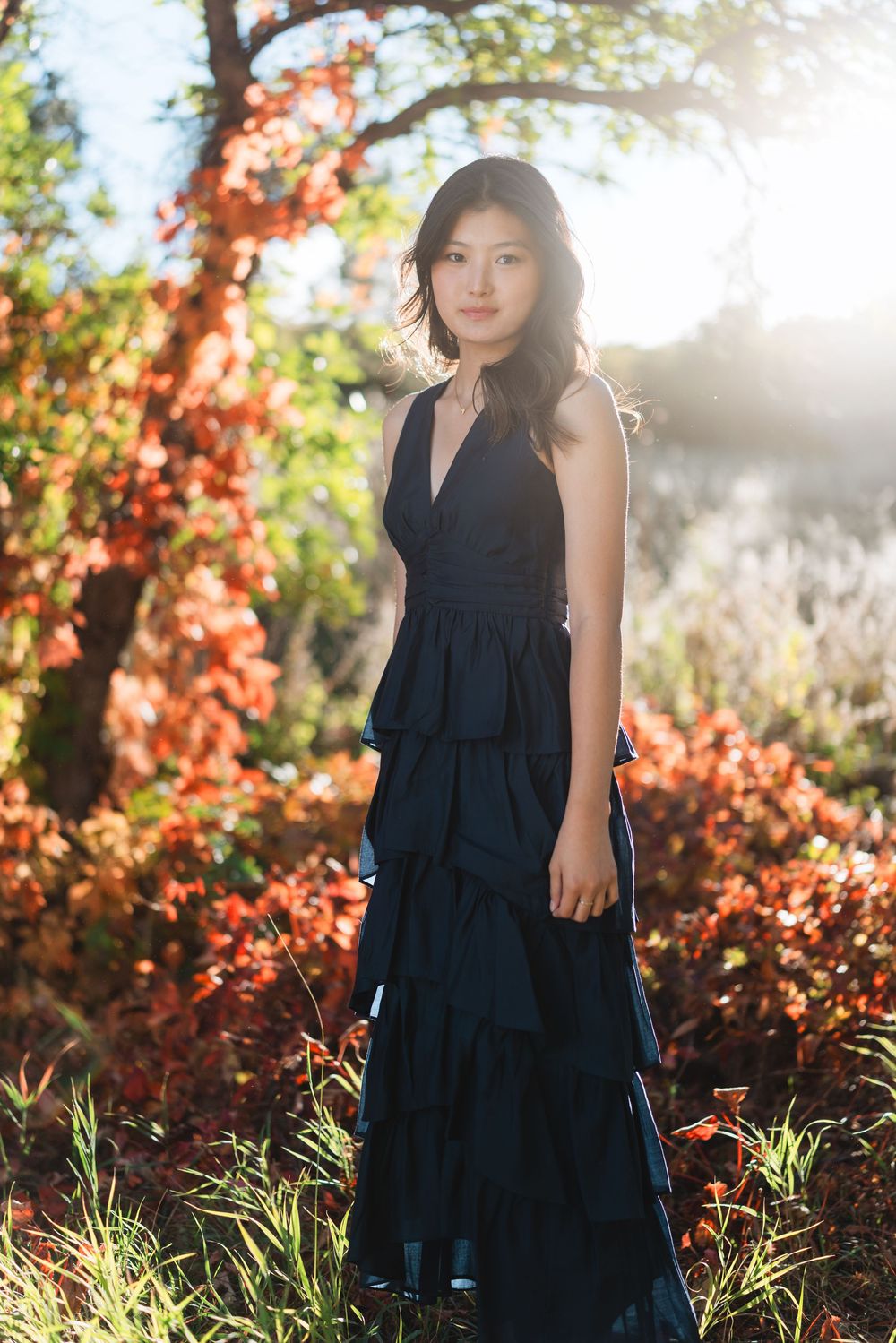 A woman in a navy blue dress poses in autumn foliage with sunlight streaming through the trees.