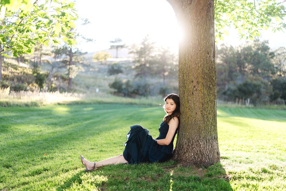 Someone sits against a large tree trunk in a sunlit grassy park on a bright summer day.