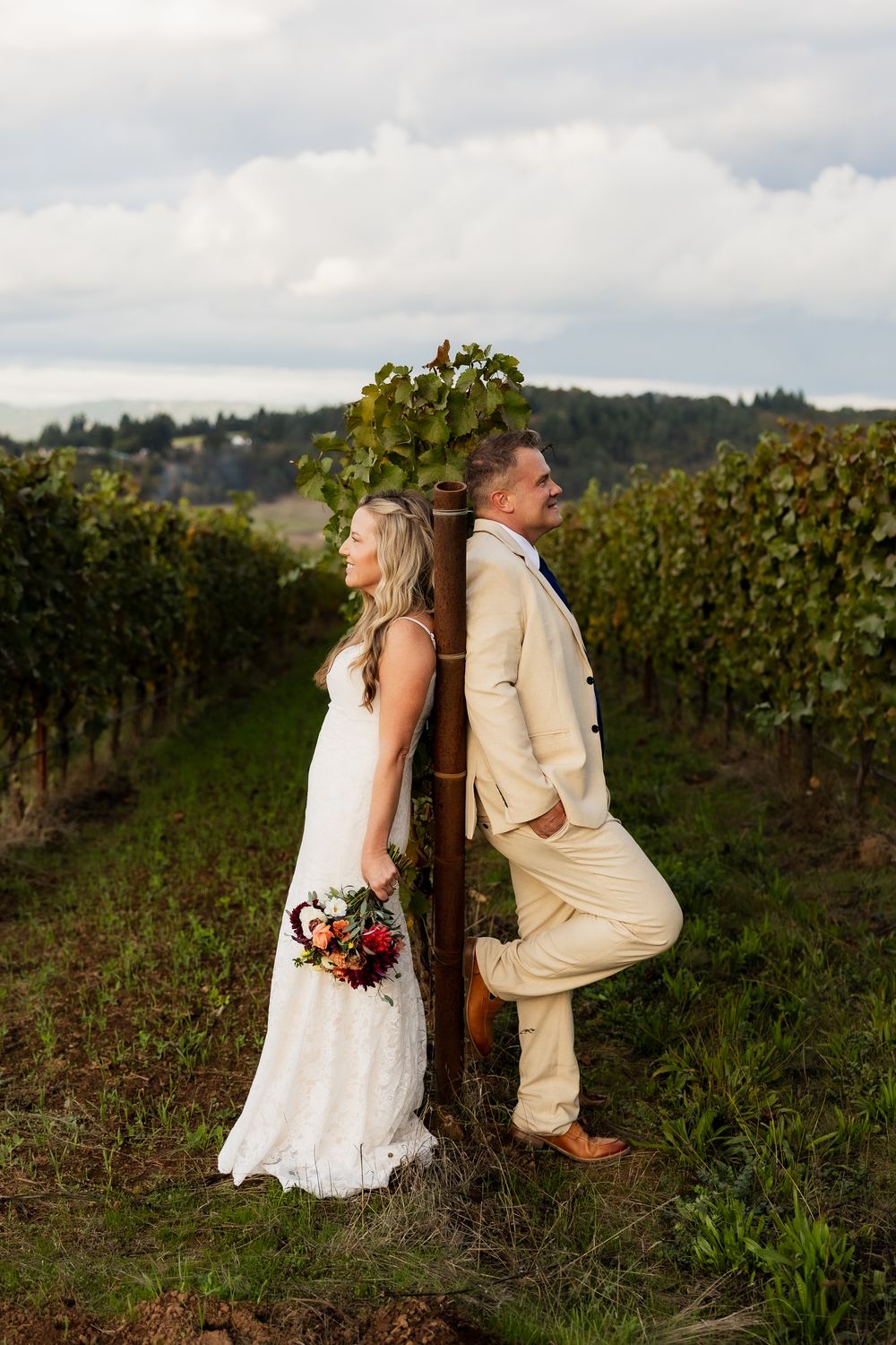 A couple poses playfully in a vineyard wearing cream and white formal attire.