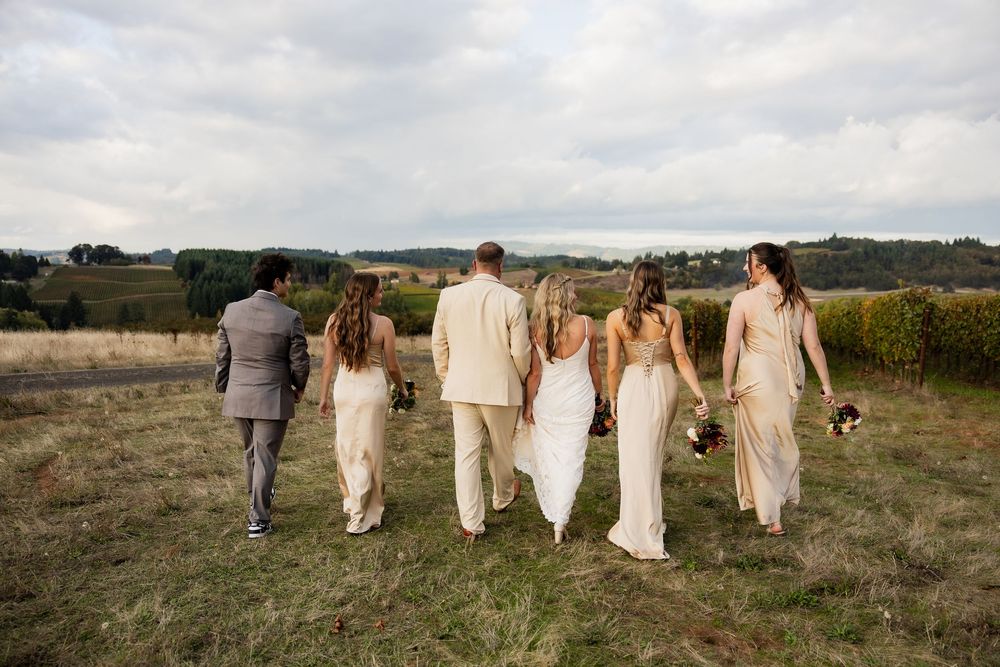 Wedding party members walk in a line across a grassy hillside under cloudy skies.