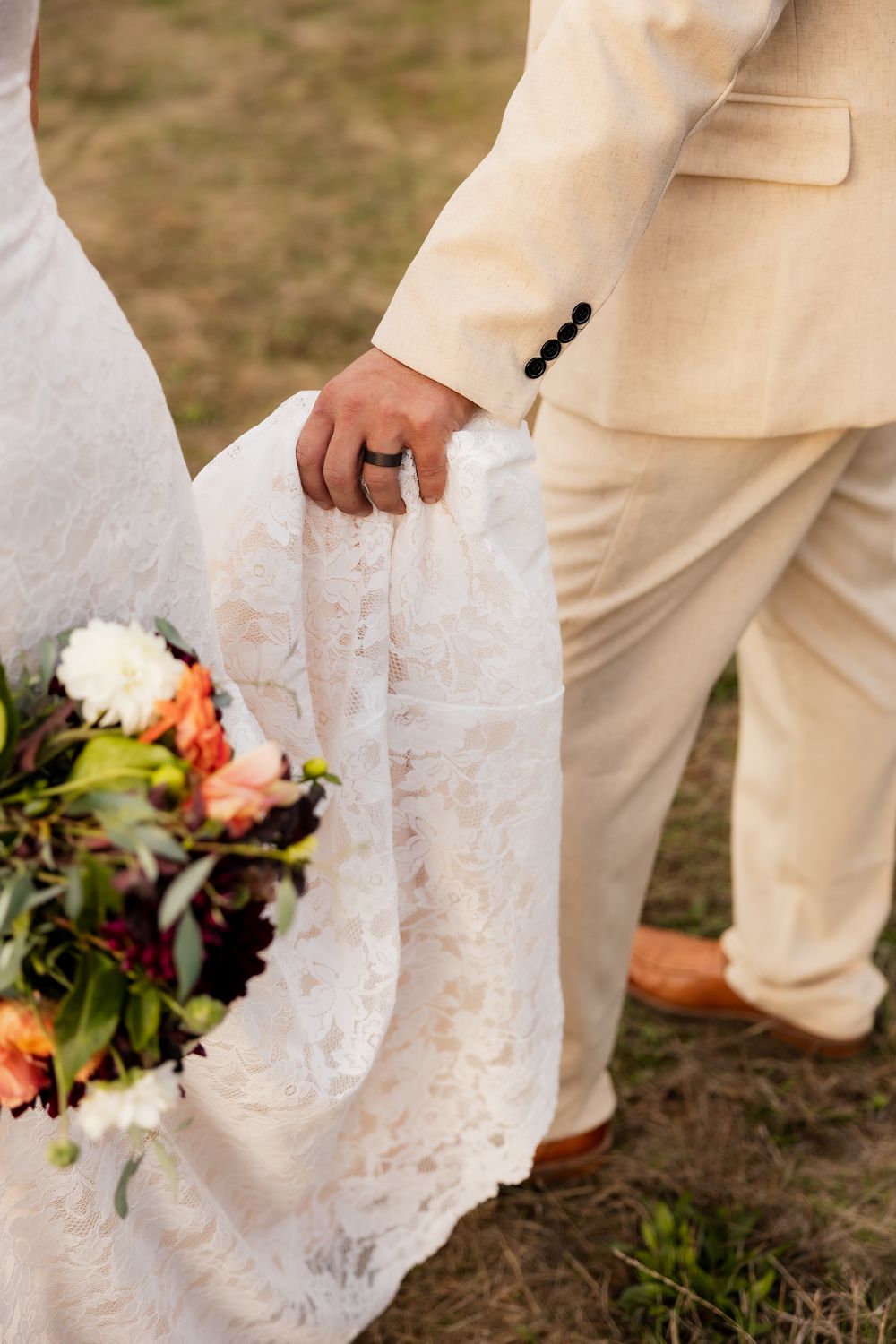 Close-up of bride in white lace dress and groom in beige suit holding autumn-colored wedding bouquet outdoors.