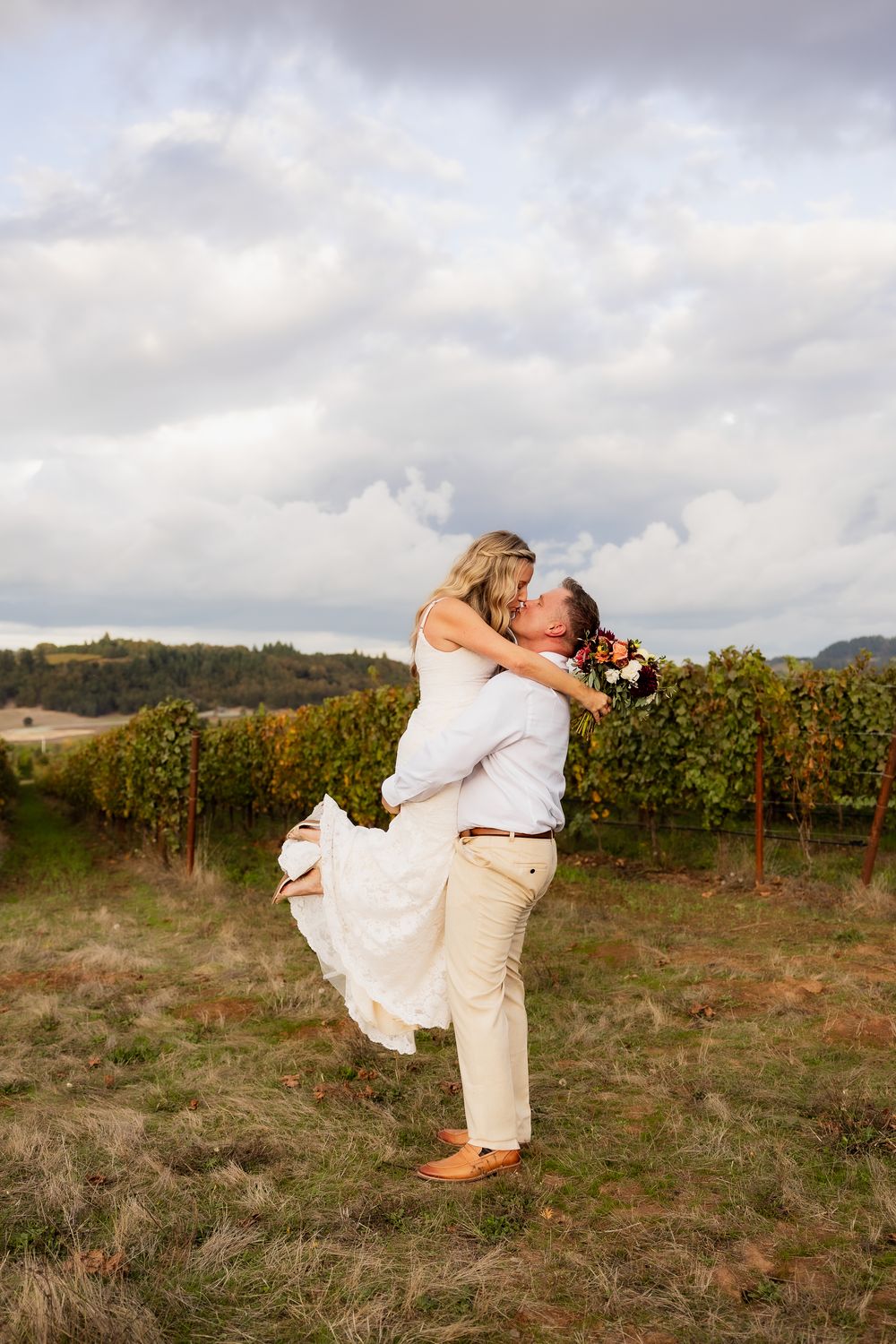 Romantic couple embraces in a scenic vineyard at sunset with golden light and rolling hills in the background.