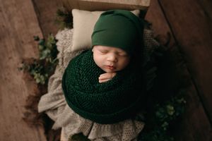 A person wearing a cozy dark green knitted beanie and scarf stands against a textured wall with climbing vines.