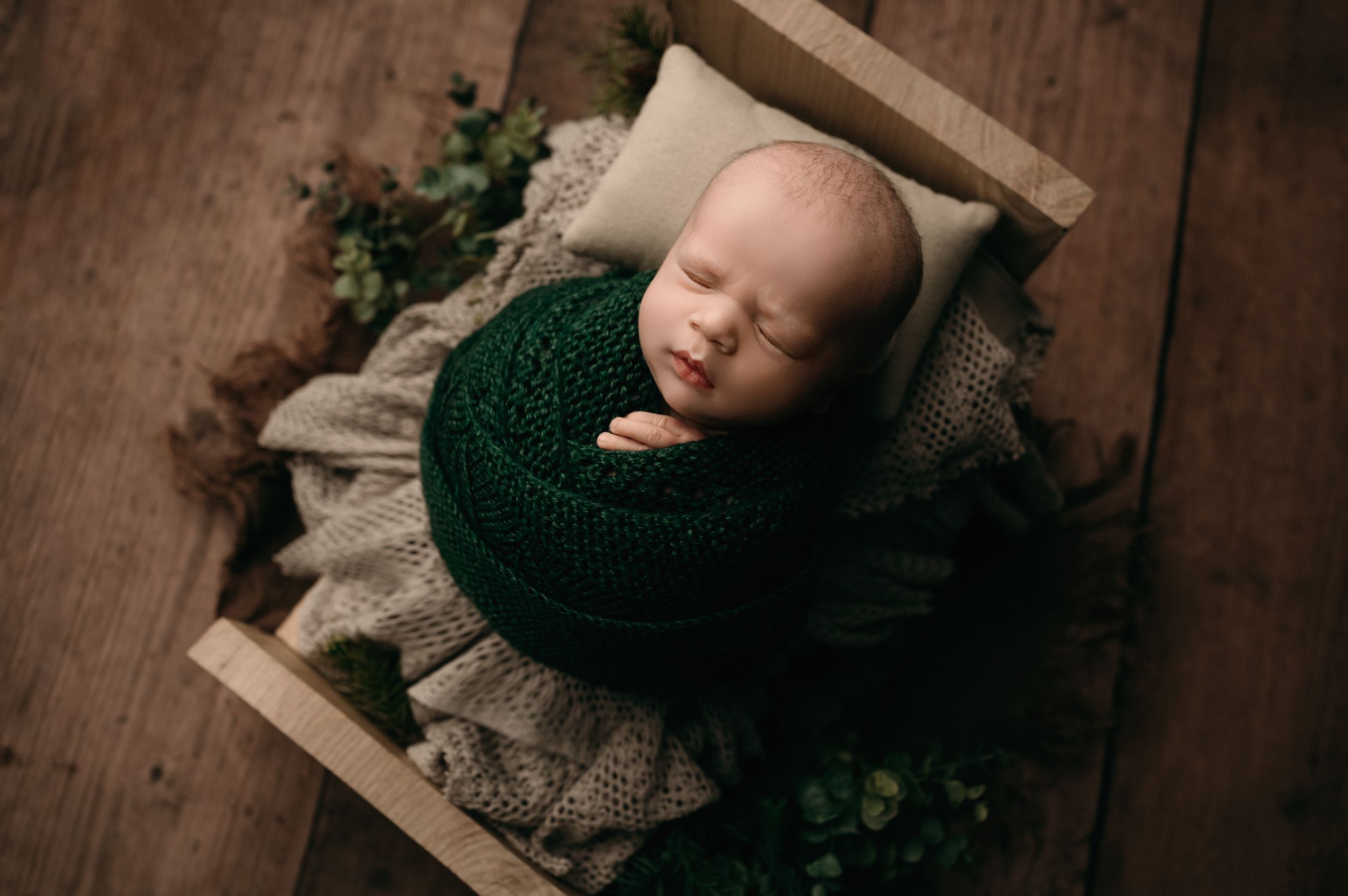 A sleeping newborn wearing a dark knit outfit rests peacefully in a rustic wooden crate on a wooden floor.