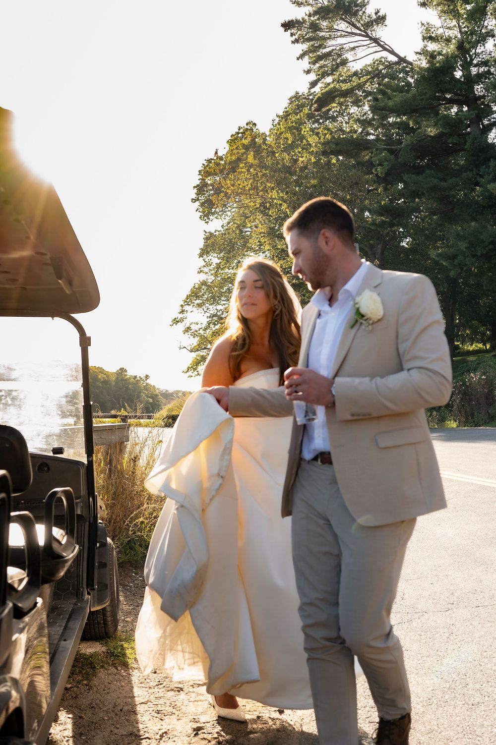 A couple in wedding attire stands near a golf cart at sunset with sunlight streaming through trees.