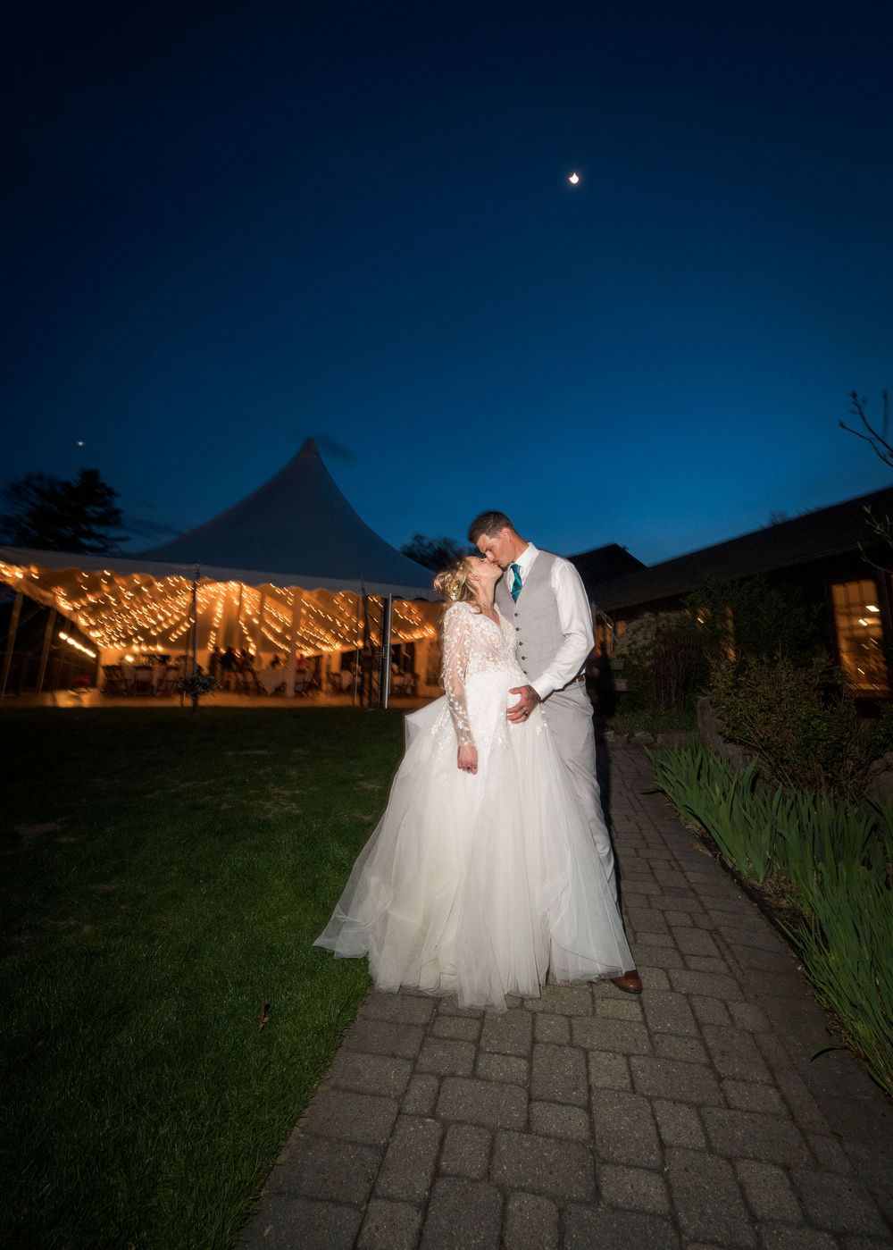 Illuminated outdoor wedding venue at dusk with stone pathway and glowing string lights overhead.