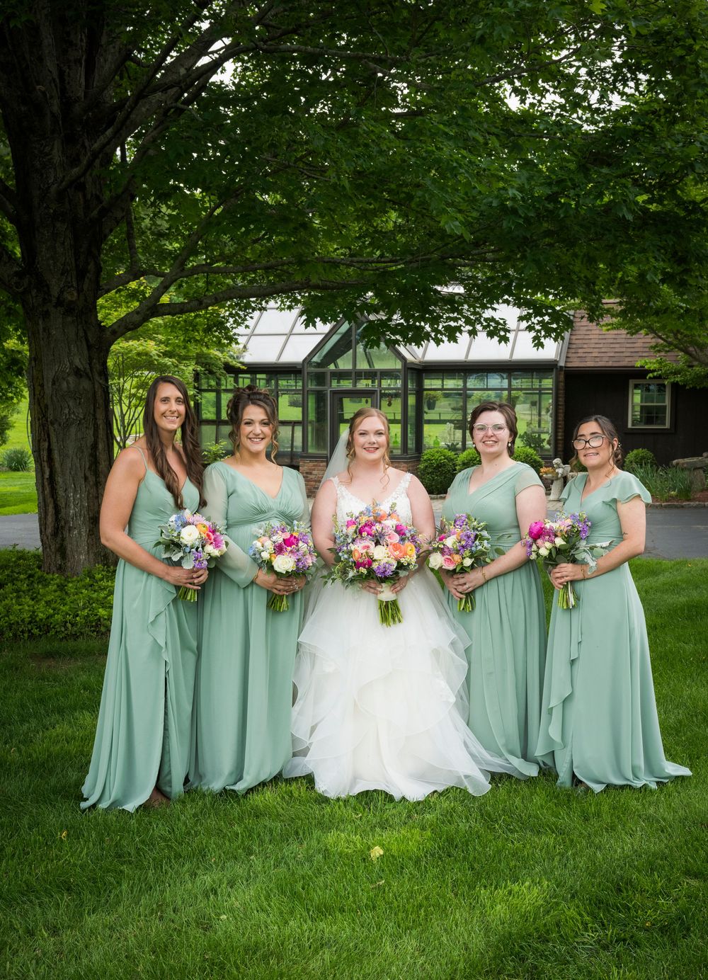 Wedding party in sage green bridesmaid dresses poses together on lush grass under trees.