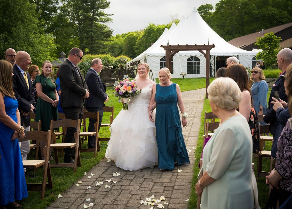Wedding ceremony guests line a stone pathway outdoors with a white tent visible in the background.
