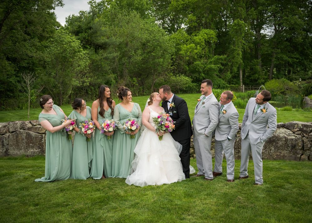Wedding party poses together on lush green lawn with trees in background during summer celebration.