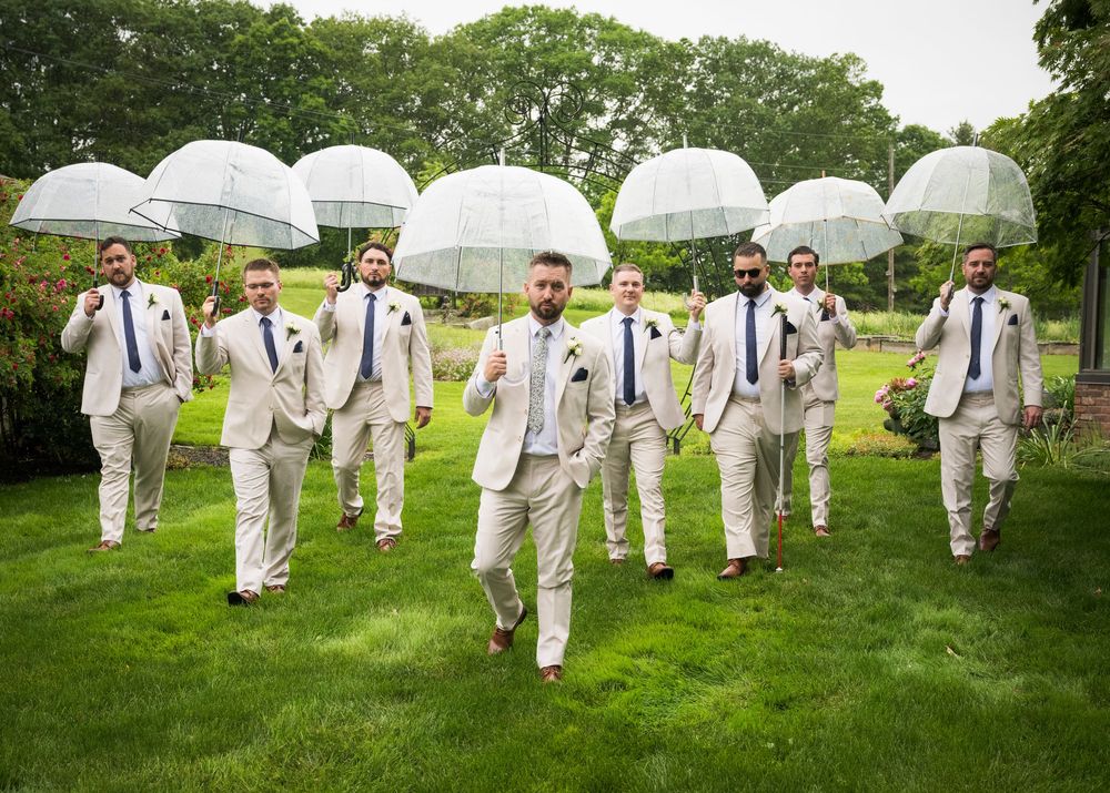 Wedding party in light colored suits jumps playfully in sync on a grassy lawn with white umbrellas on a cloudy day.