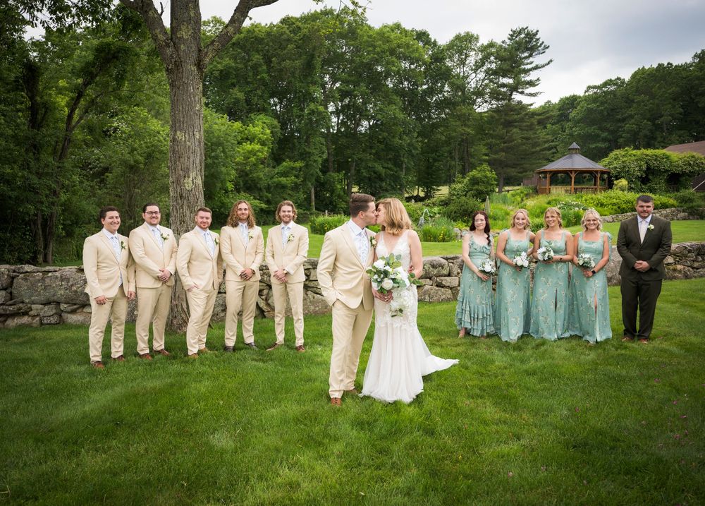 Wedding party poses together on lush green lawn with gazebo and stone wall in background.