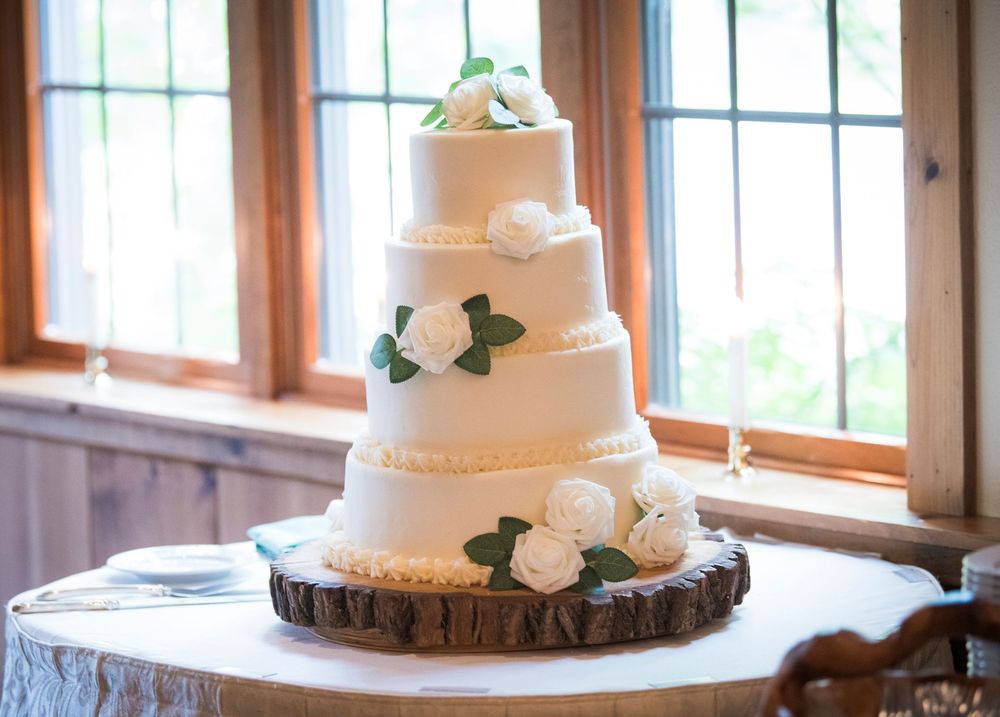 A rustic wedding cake display featuring multiple white tiered cakes decorated with greenery on wooden stands near windows.