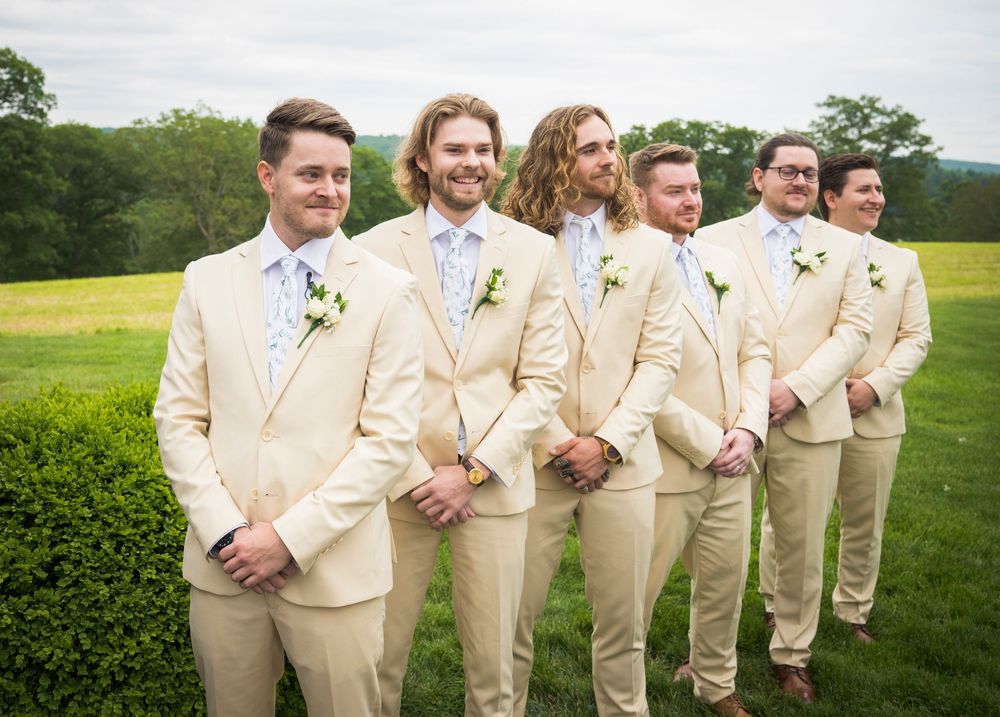 Wedding party members in tan suits stand together on a lush green lawn during an outdoor celebration.