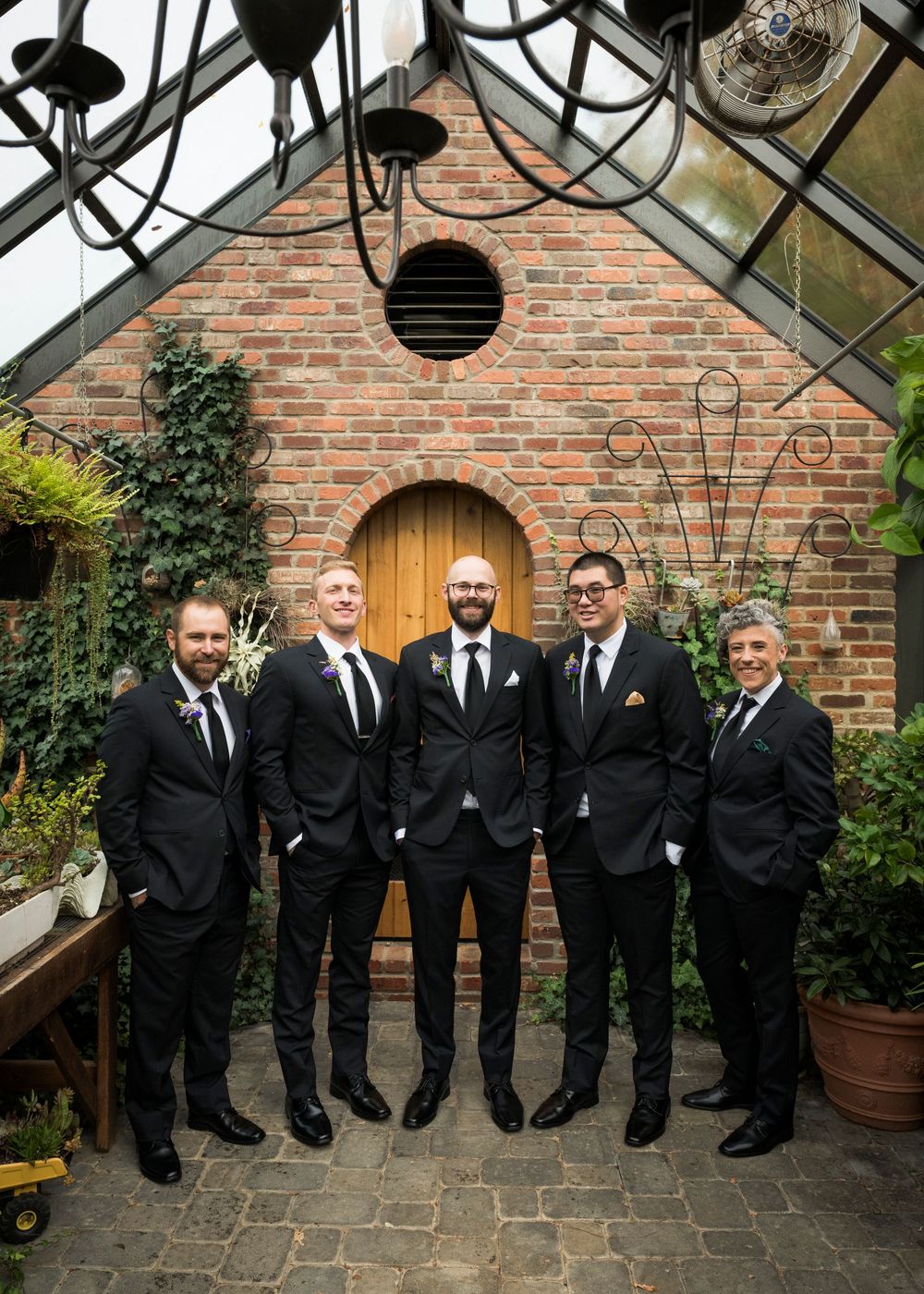 Groomsmen in black suits and ties pose together in front of a brick wall and greenhouse structure.