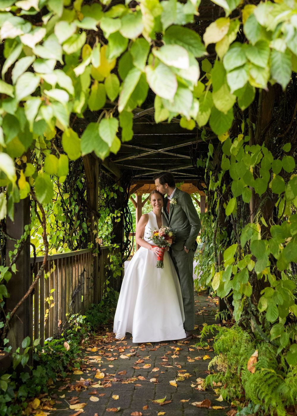 A romantic wedding couple poses under a lush green vine-covered arbor with scattered autumn leaves on the ground.