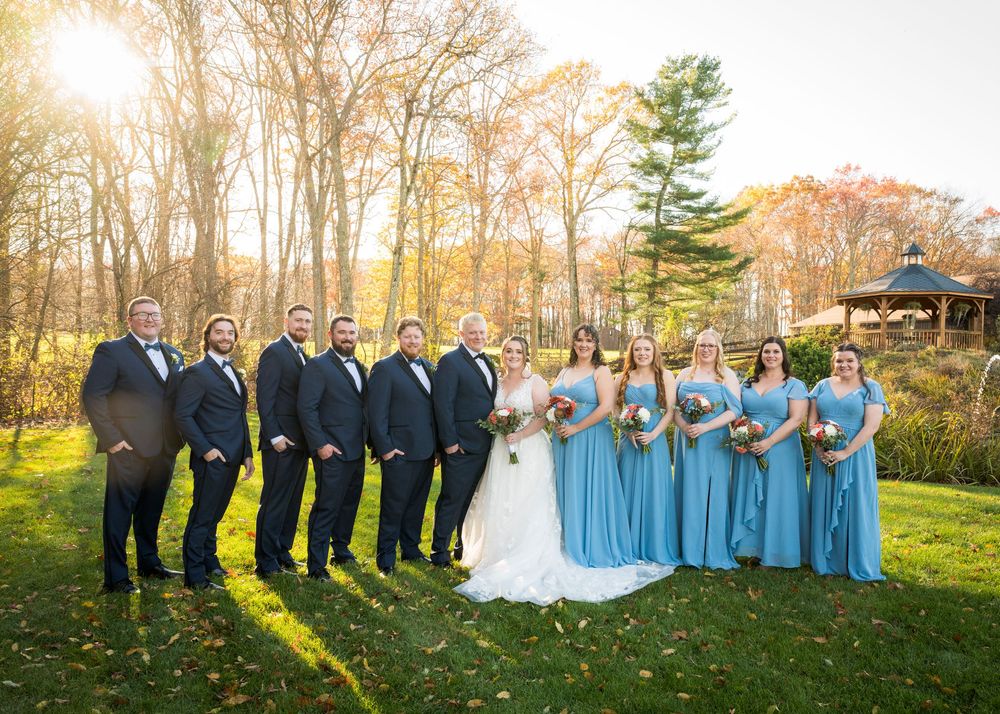 Wedding party in navy suits and light blue dresses poses together on autumn lawn at sunset.