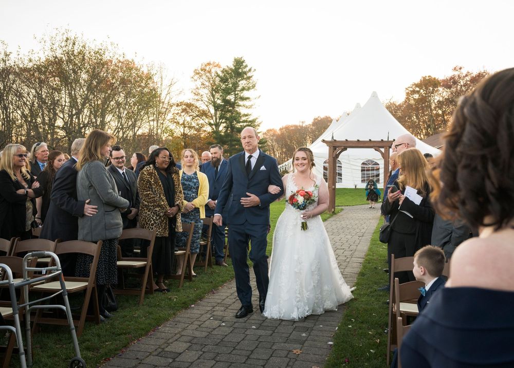 Outdoor wedding ceremony procession along a stone path with guests seated on both sides at sunset.