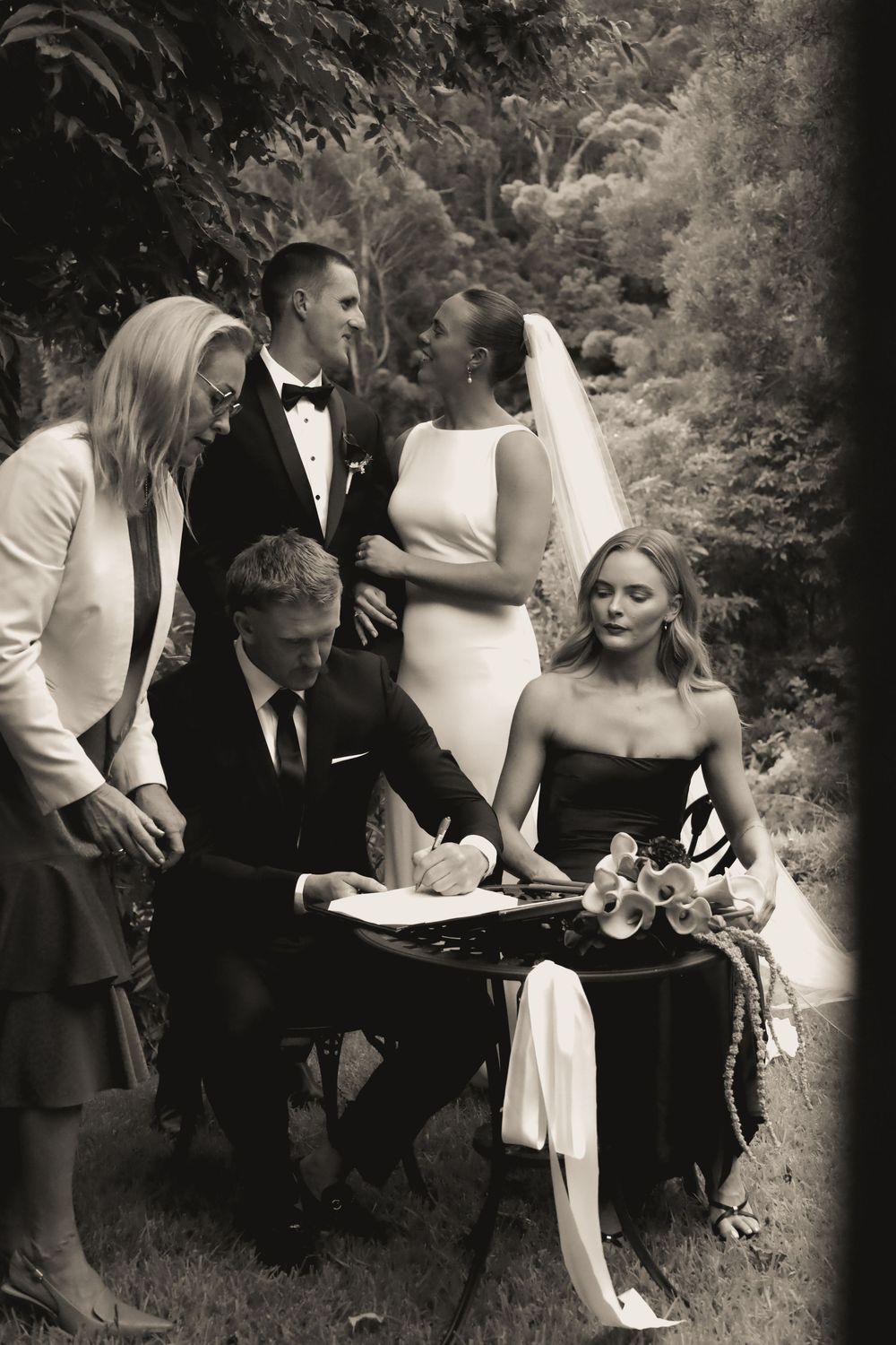 A series of black and white wedding photos showing a couple signing documents in an outdoor garden setting.