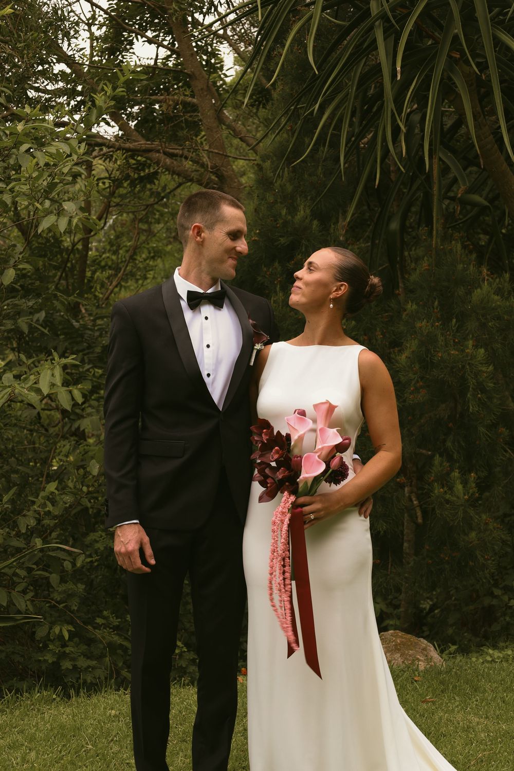 A couple in wedding attire stands together in a lush garden setting with tropical plants in the background.