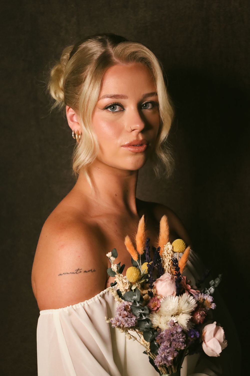 Portrait of person in off-shoulder white dress holding colorful dried flower bouquet against dark background.