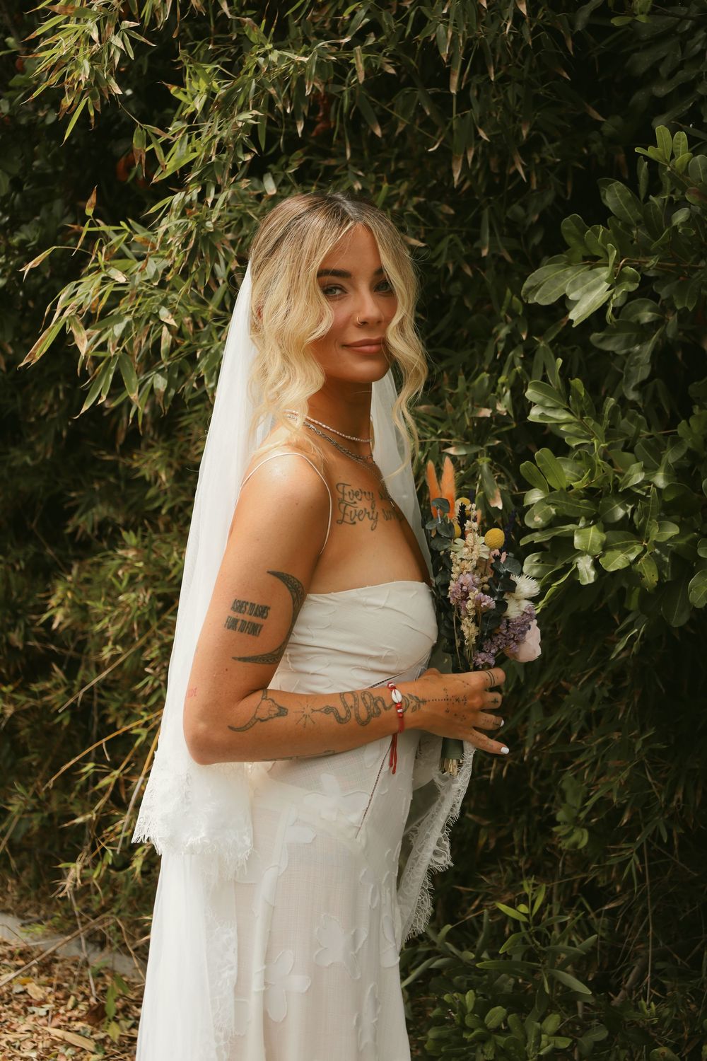 A bride in a white strapless wedding dress and veil poses in front of lush greenery, showing delicate tattoos.