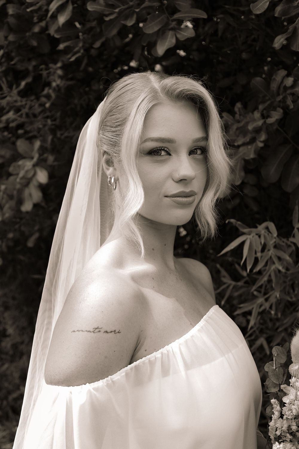 Black and white portrait of a bride wearing an off-shoulder dress and veil against a leafy background.
