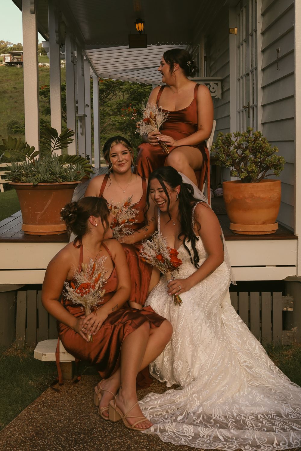 A group in rust-colored bridesmaid dresses and white wedding gown share candid laughing moments on a porch.