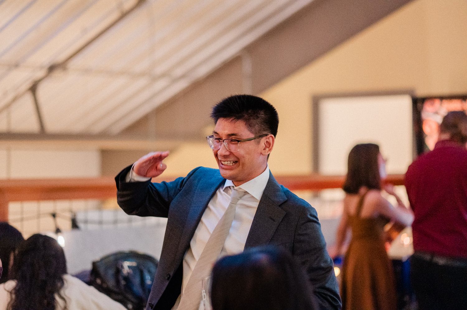 A groom wearing glasses and a gray suit raises their hand during wedding reception indoors