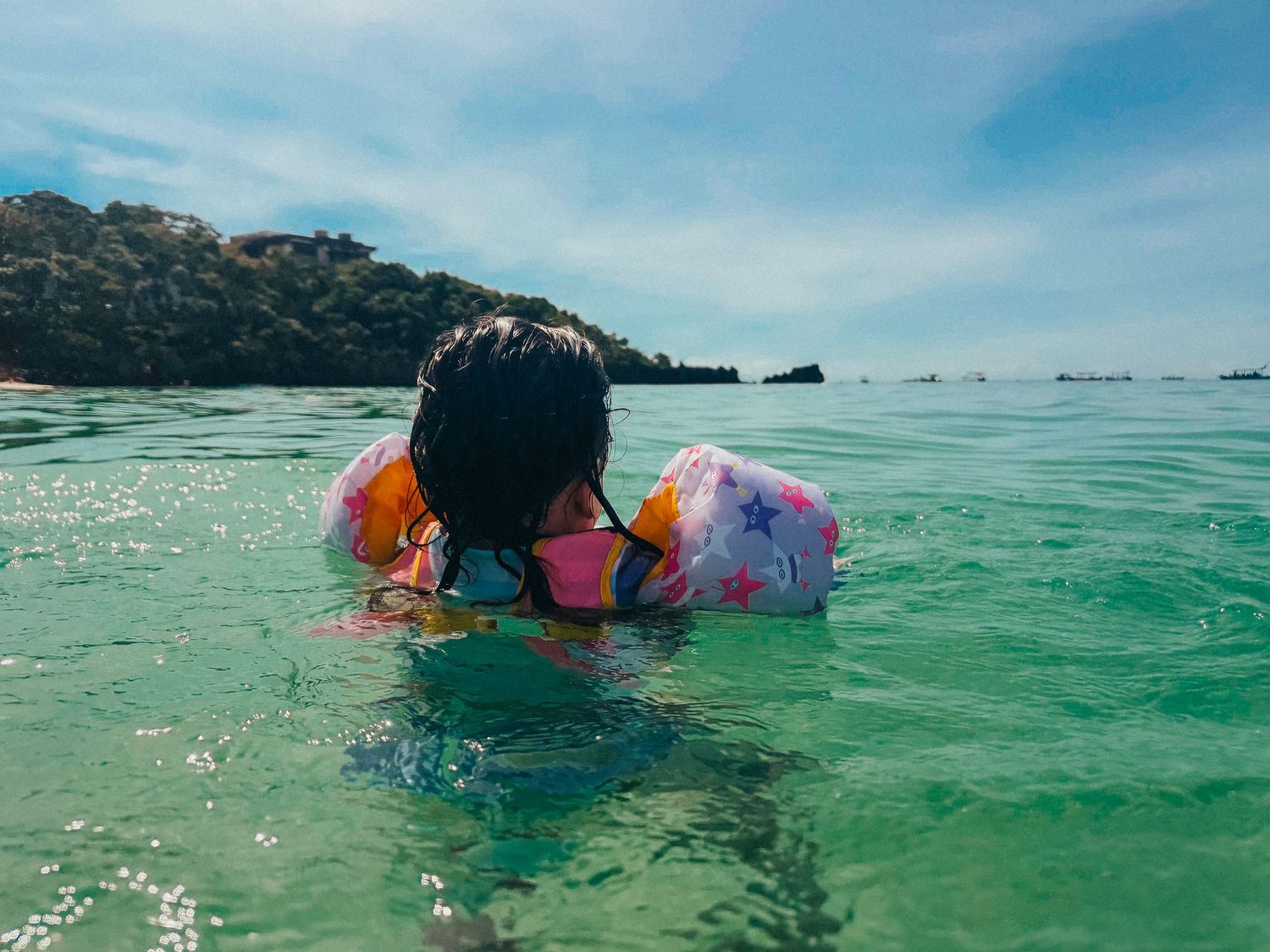 Someone wearing floaties swims in turquoise waters near a tropical island with boats visible in the distance.