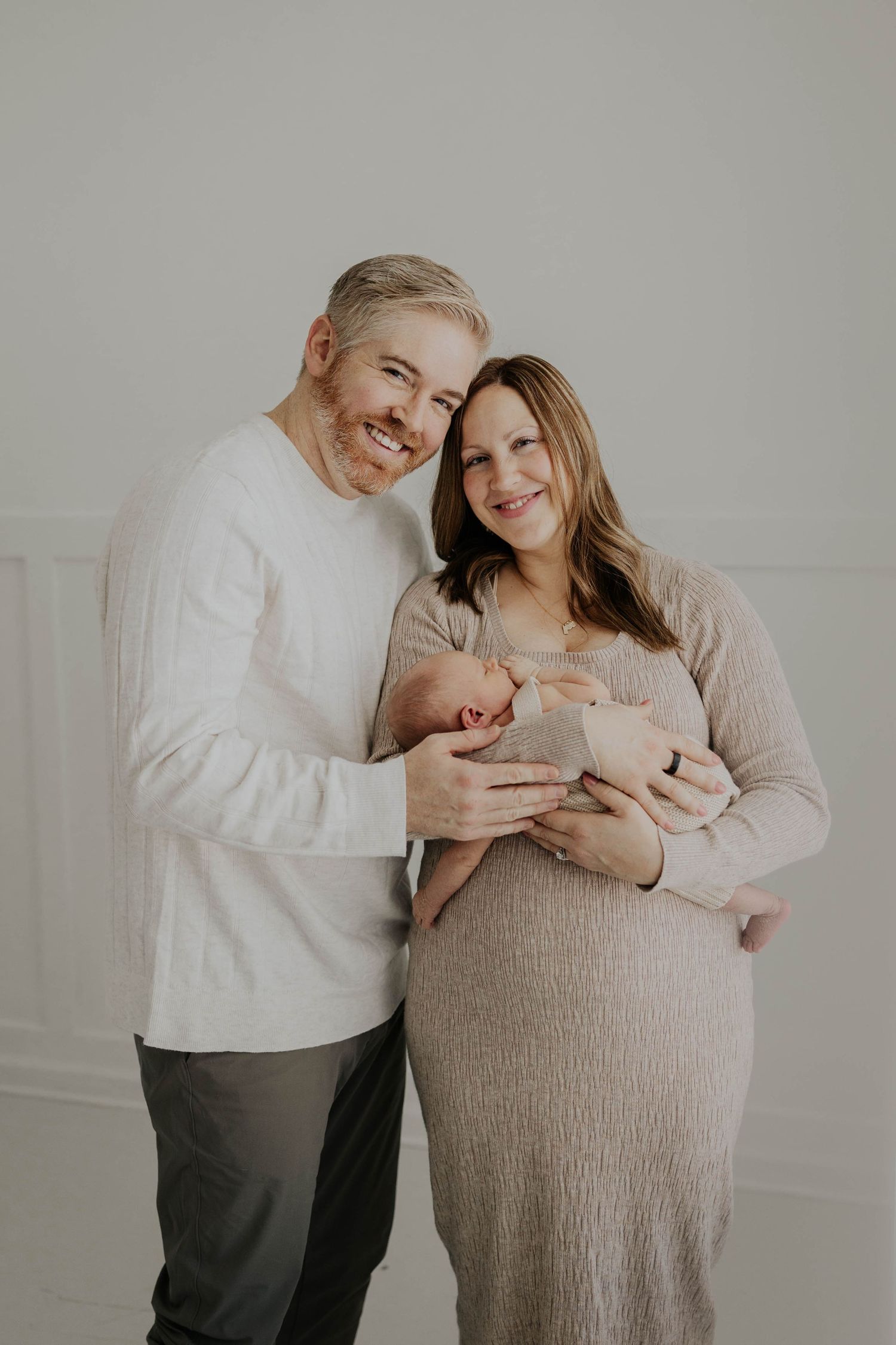 A couple in light neutral colors share tender moments during a maternity photo session against a light gray backdrop.