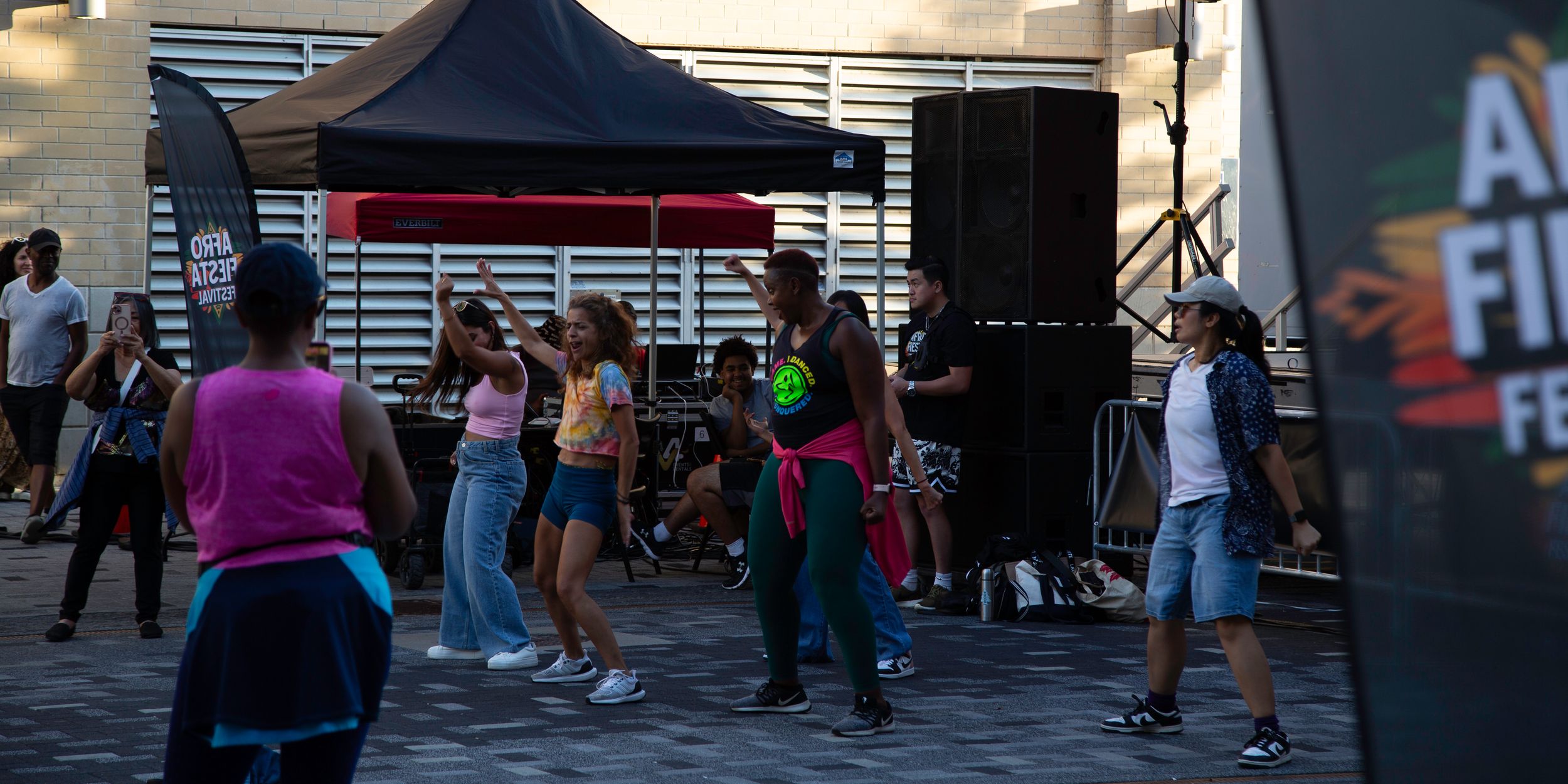 People dance together on a paved outdoor plaza near a black canopy tent and speakers at a lively community festival.