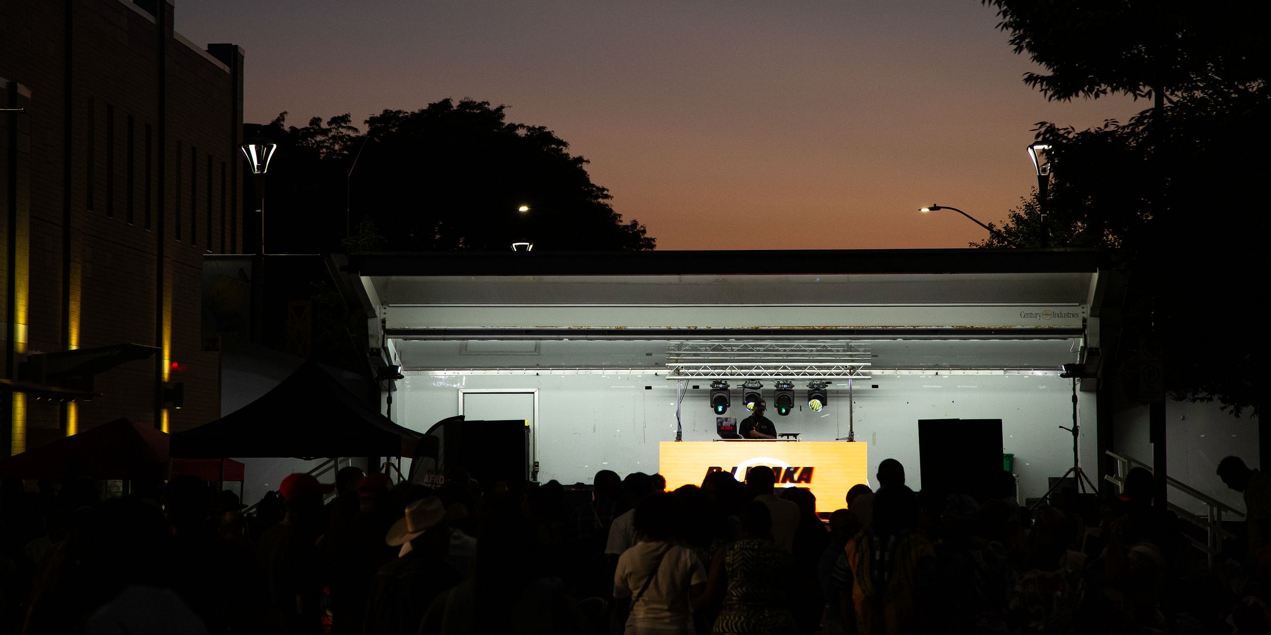 Crowd silhouetted at dusk watching a DJ perform on a lit outdoor stage under an orange and purple twilight sky.