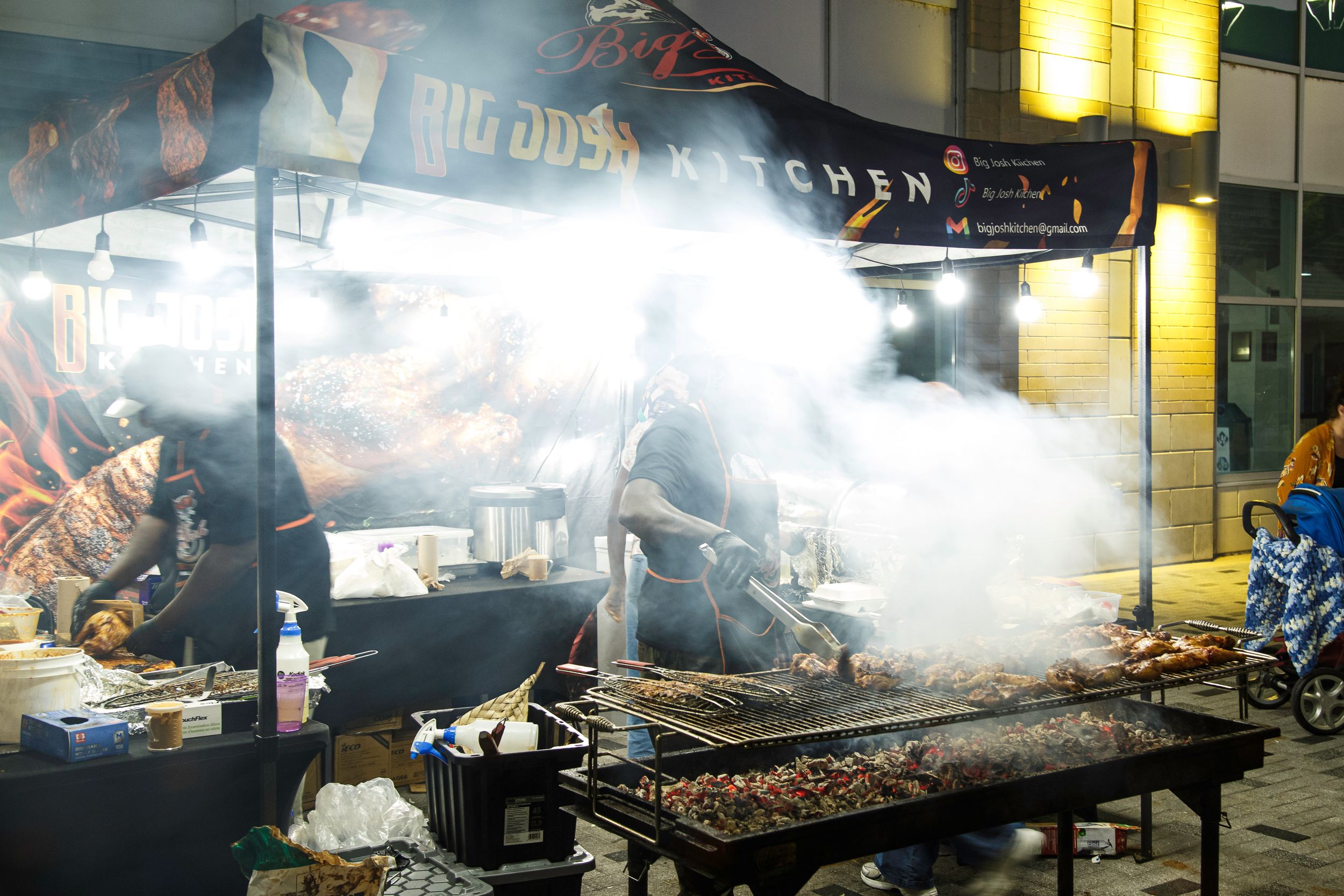 Street food vendors grill skewered meats over a smoking open barbecue beneath a kitchen banner at a lively outdoor night market.
