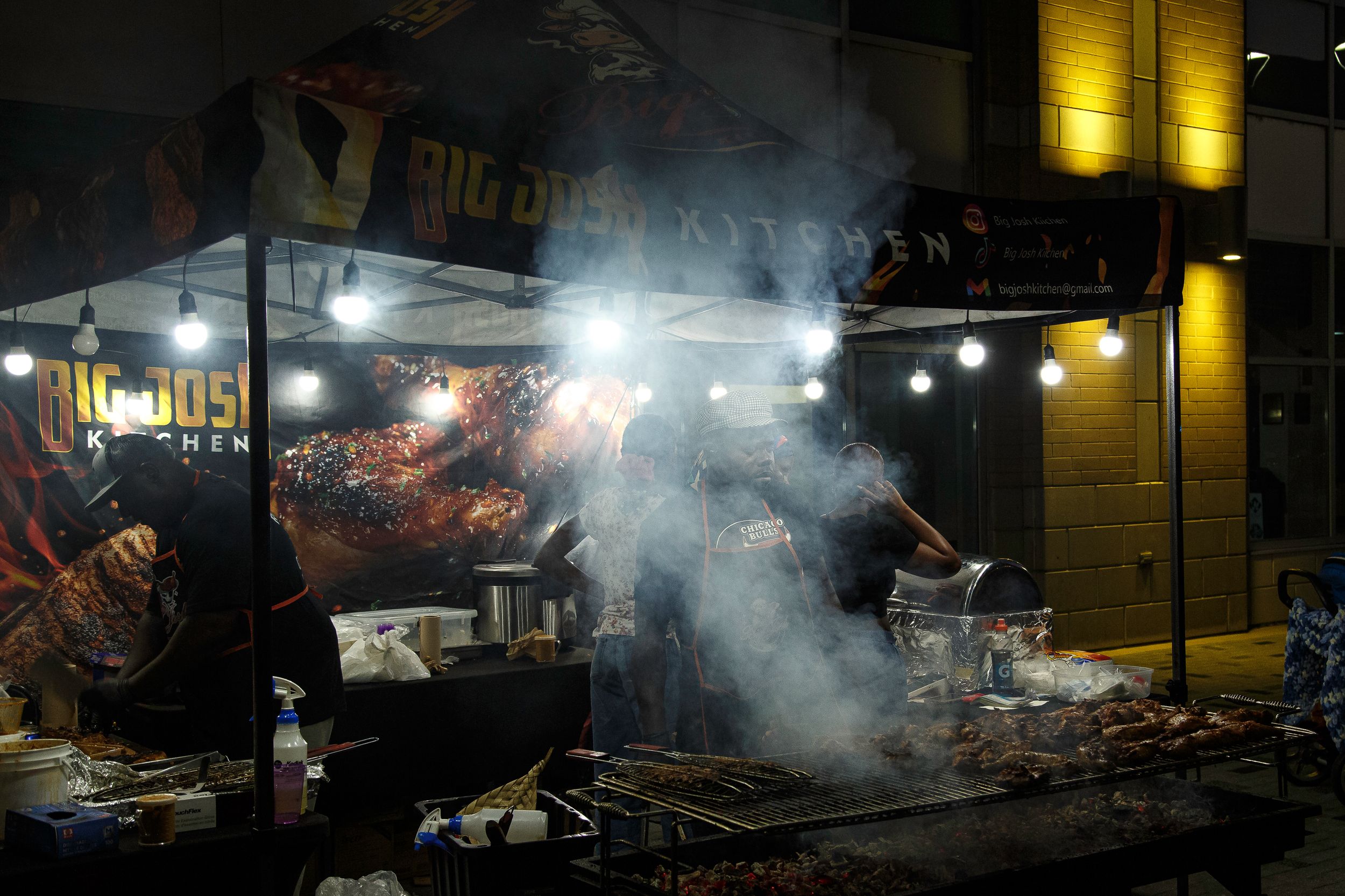 Smoke rises from a nighttime street food stall with string lights, a grill and displays of roasted meats beneath a banner.