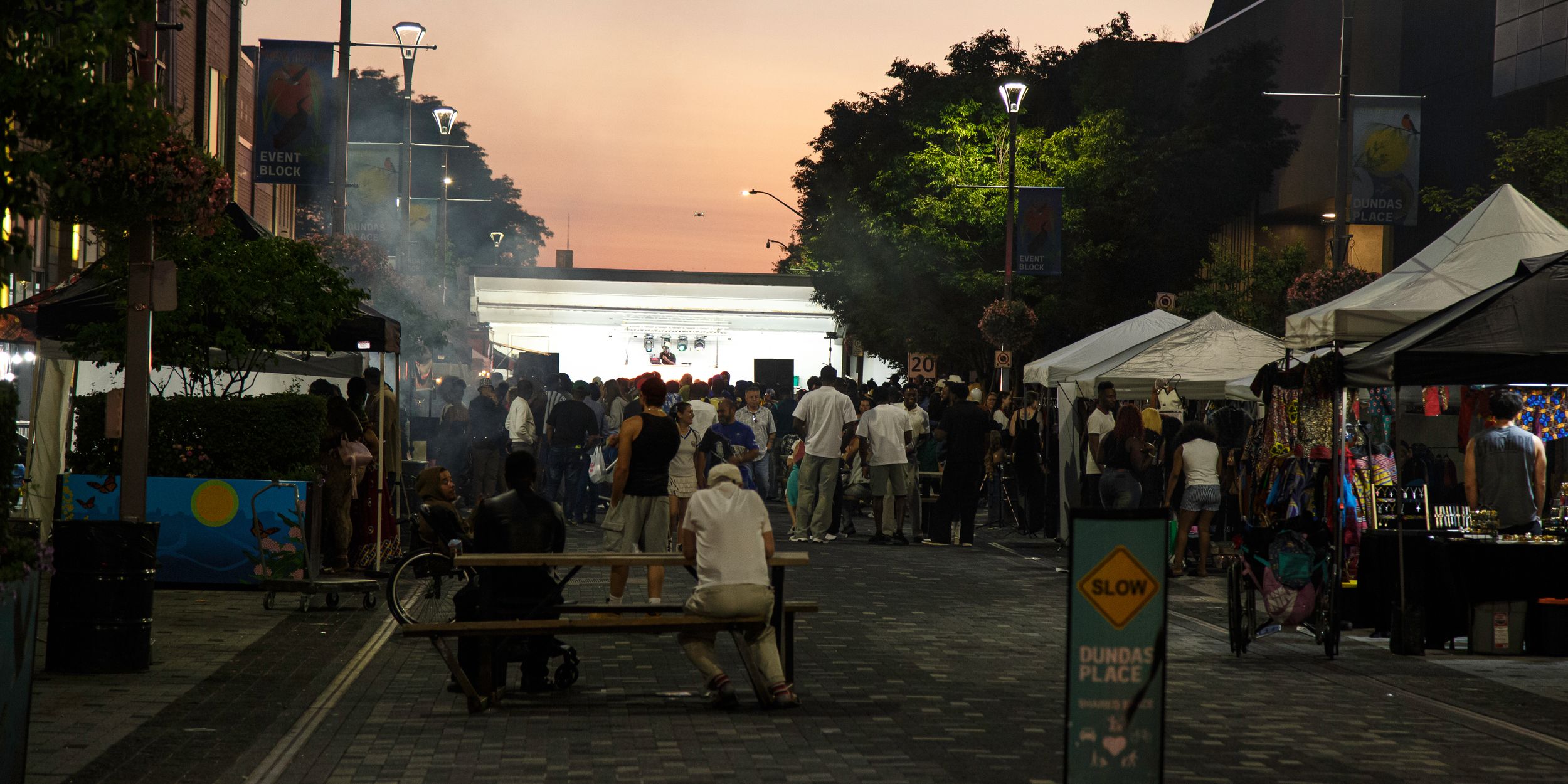 Crowds gather at an outdoor evening market with vendor tents, drifting smoke, and a glowing building under a pink dusk sky.